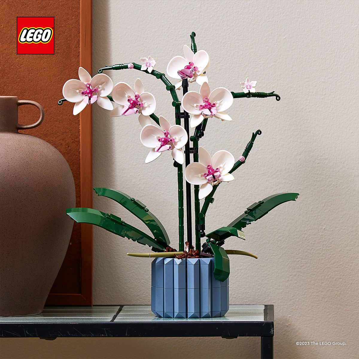 LEGO Orchidee mit weißen Blüten und pinkfarbenen Blütenstempeln in blauem, achteckigem Topf auf schwarzem Tisch vor weißer Wand und braunem Gefäß.