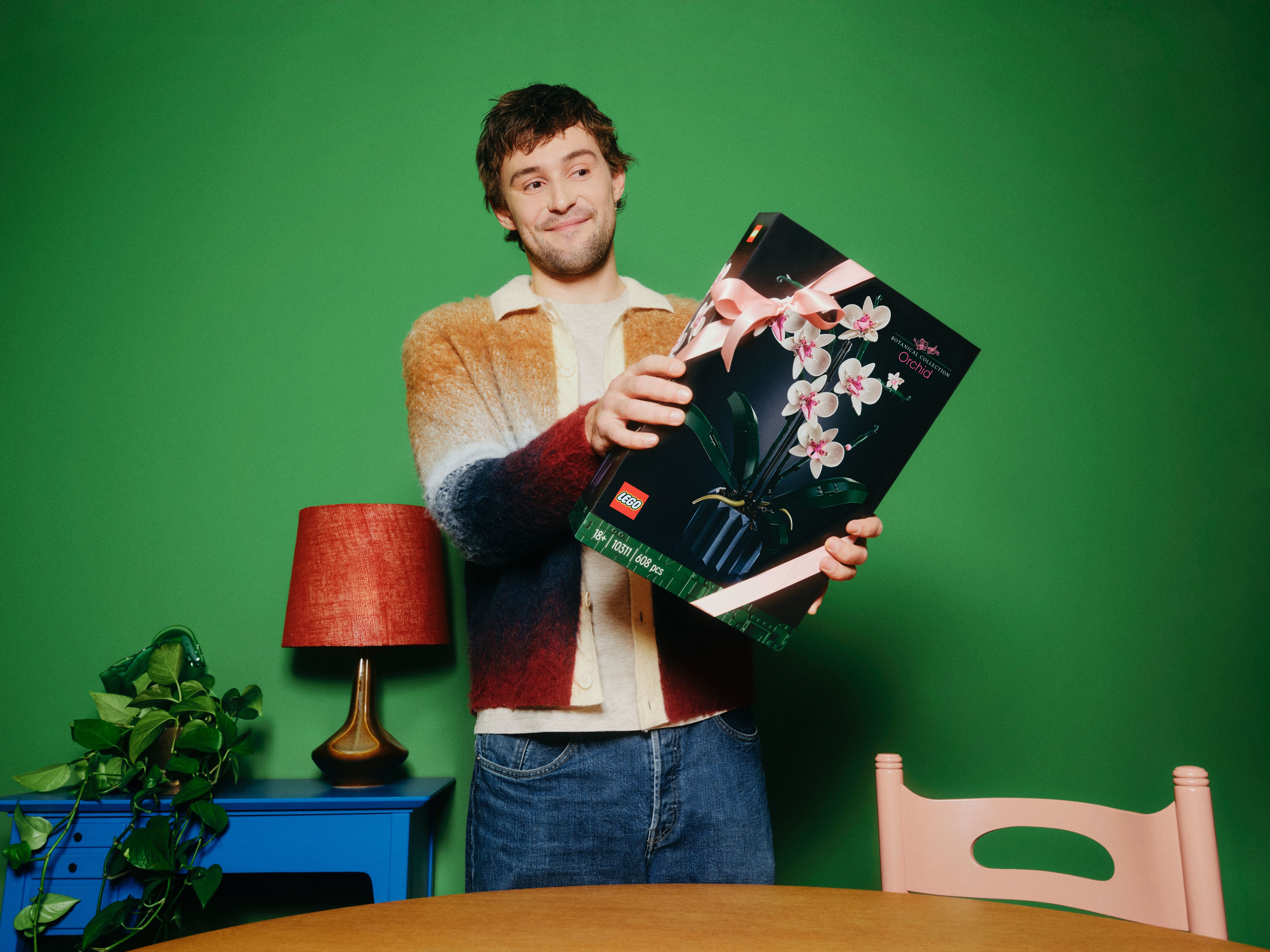 Person hält eine LEGO Orchidee-Bausatzbox mit rosa Orchideenblüten vor grünem Hintergrund, neben blauem Tisch mit Pflanze und roter Lampe.