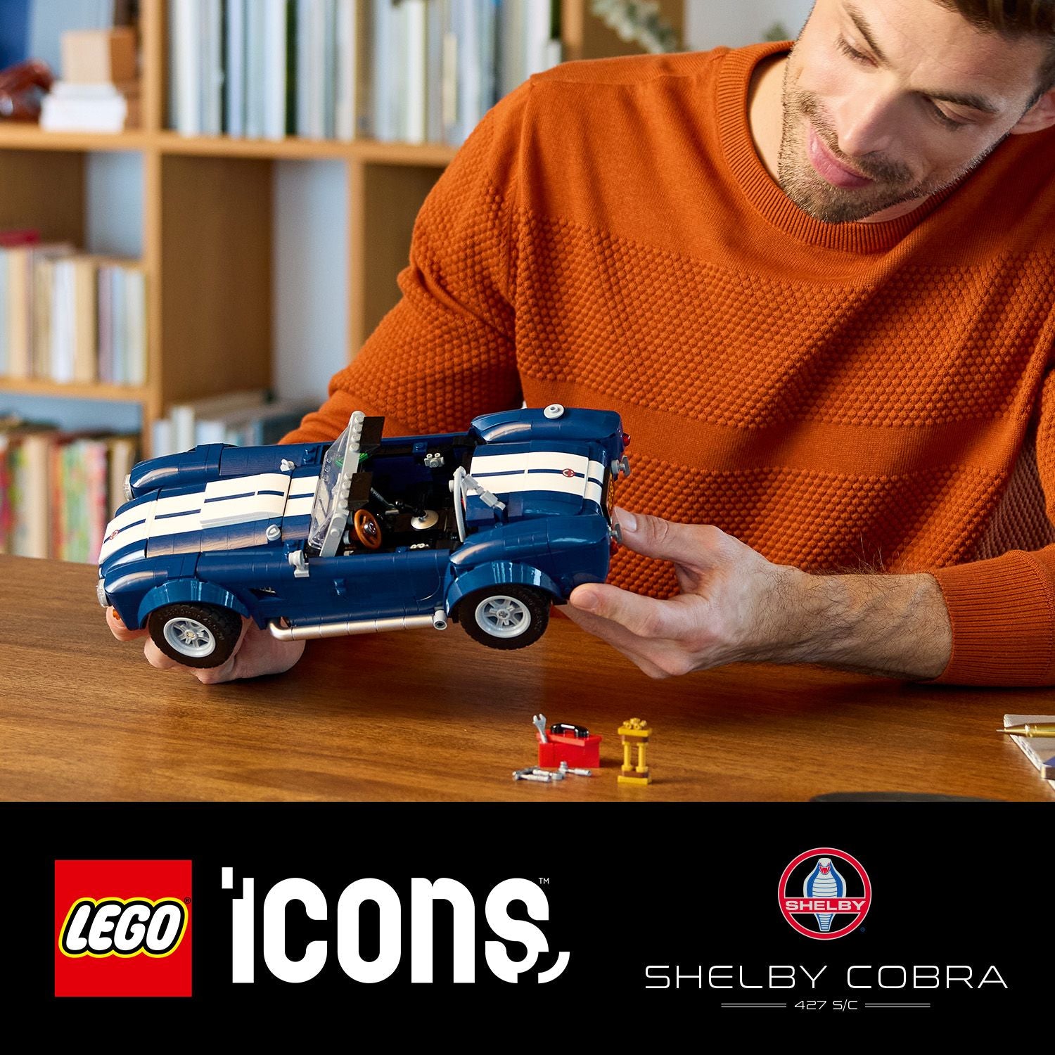 Person hält ein blau-weiß gestreiftes LEGO Modell des Shelby Cobra 427 S/C auf einem Holztisch, daneben LEGO Werkzeug und ein gelbes Objekt.