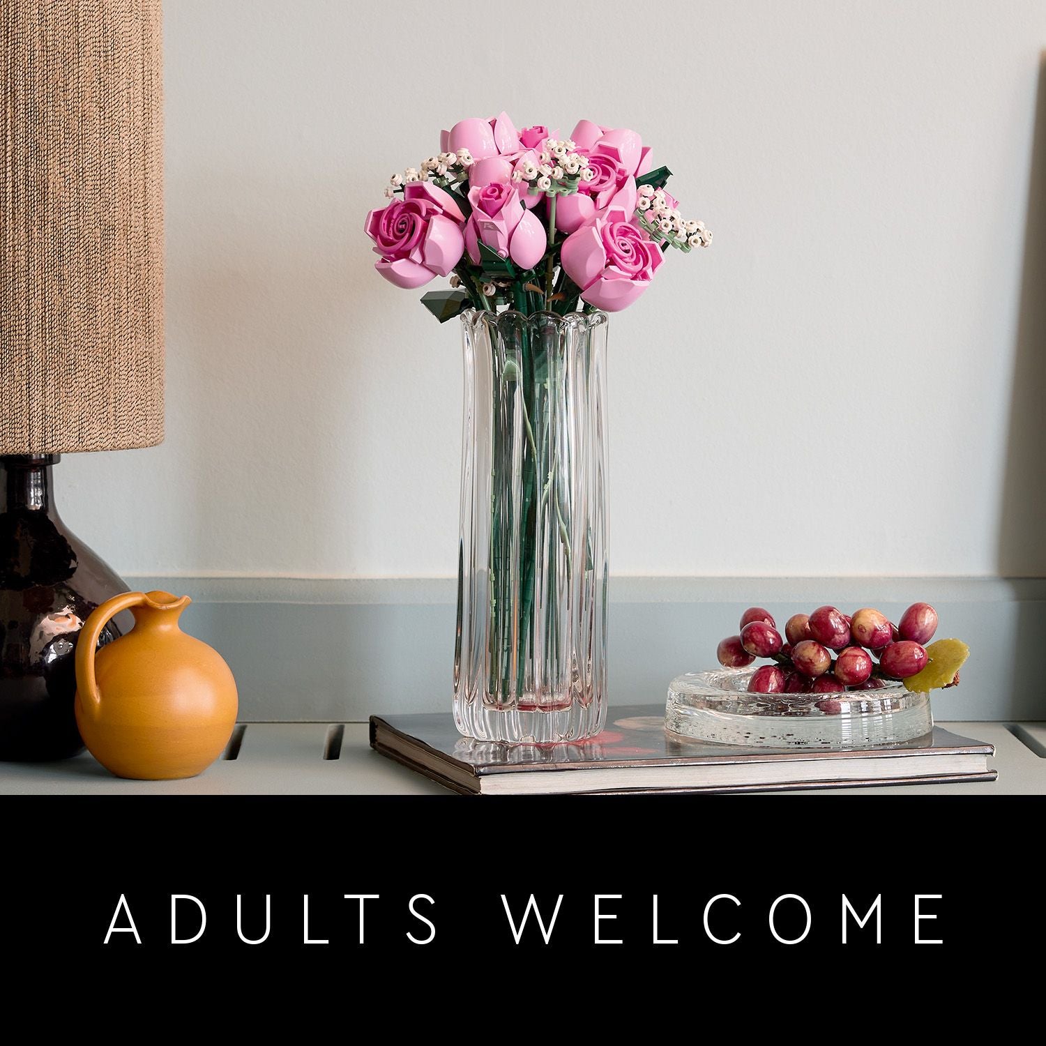 Vase mit rosa Rosen und Schleierkraut auf einem Buch, daneben eine gelbe Keramikkanne und eine Glasschale mit roten Trauben auf einem grauen Tisch, darunter der Schriftzug 'ADULTS WELCOME'.