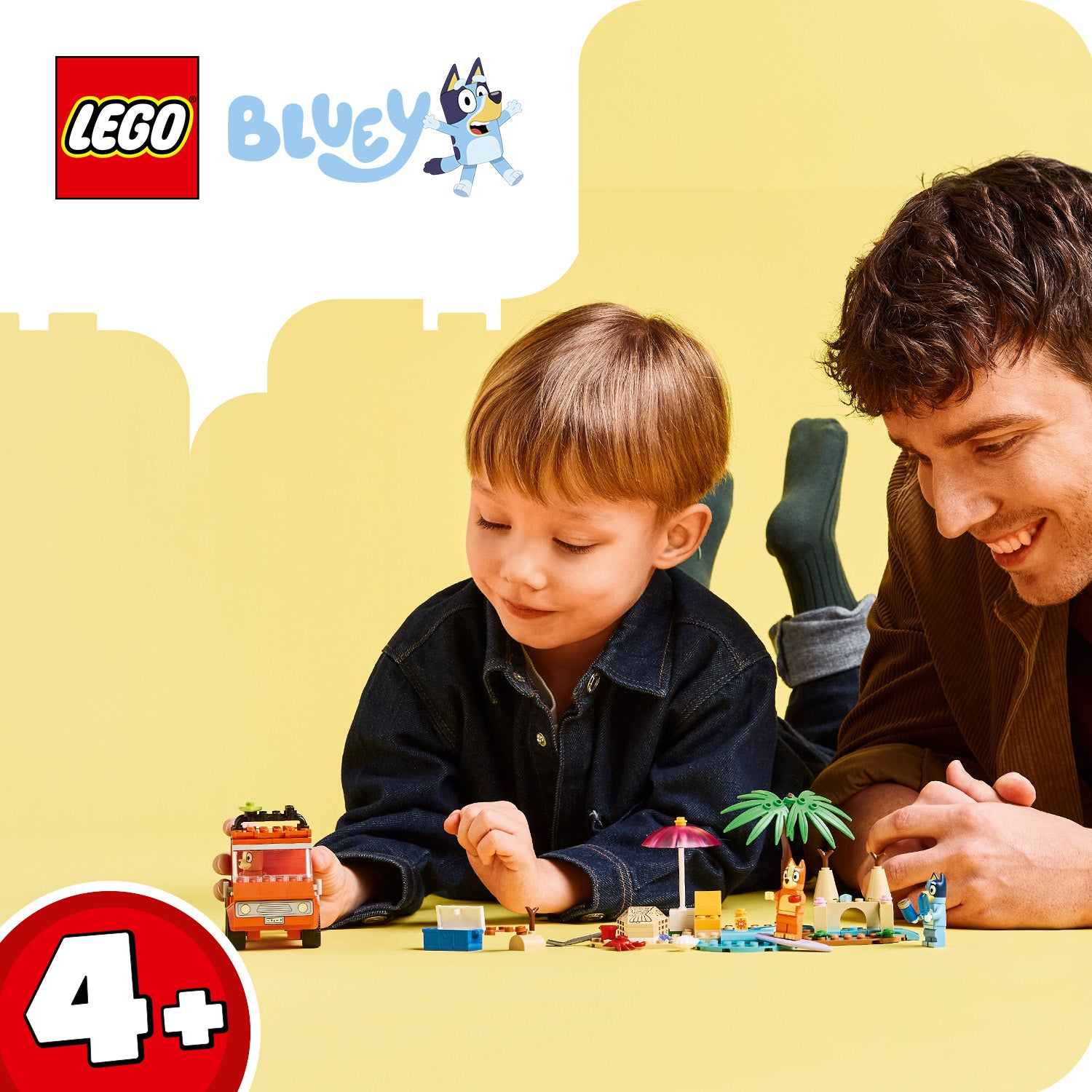 Kind und Erwachsener liegen auf dem Boden und spielen mit LEGO Bluey Strandspielzeug, darunter ein rotes Fahrzeug, eine Palme und kleine Figuren.
