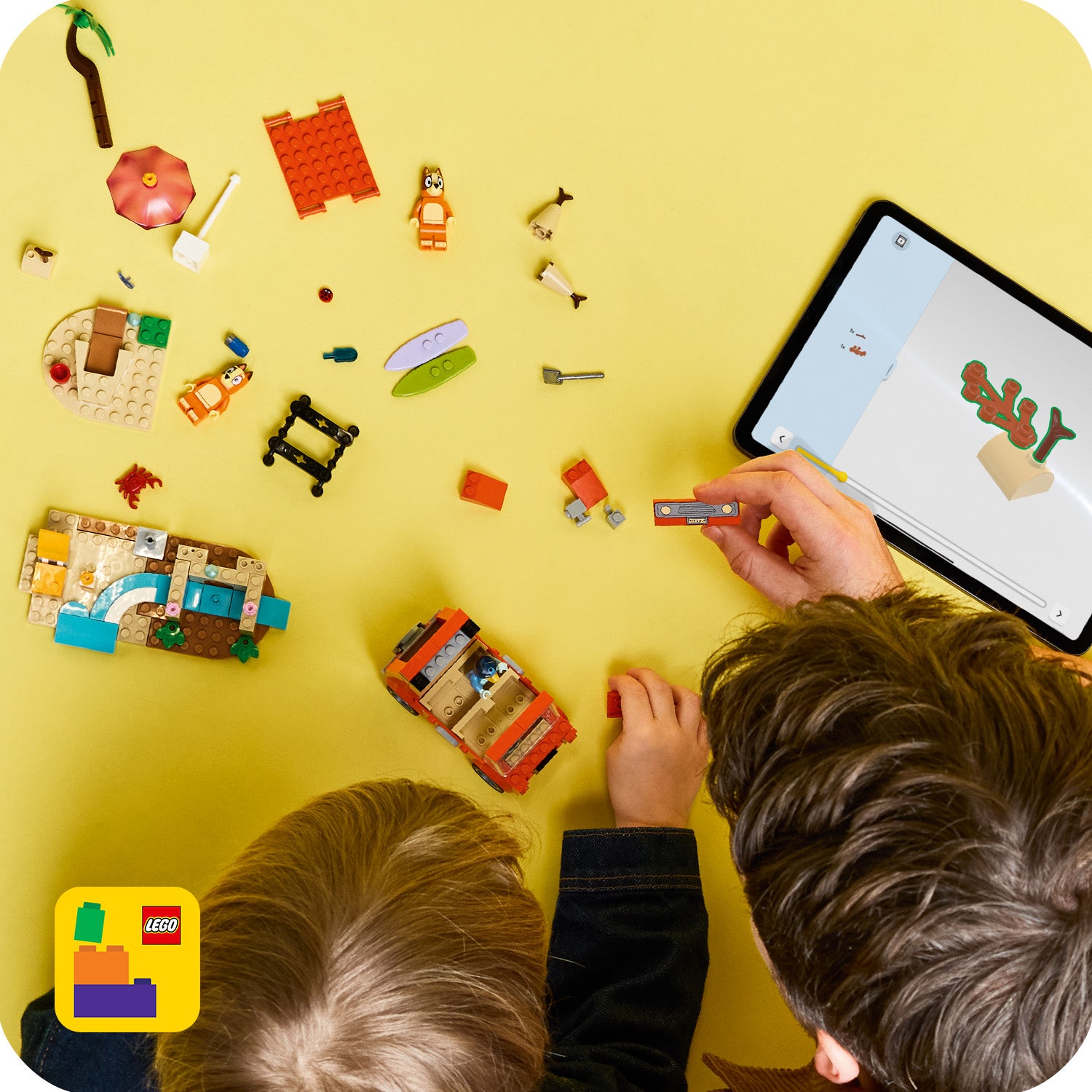Zwei Kinder bauen mit LEGO Steinen auf gelbem Untergrund, daneben ein Tablet mit digitaler Bauanleitung für LEGO Bluey.