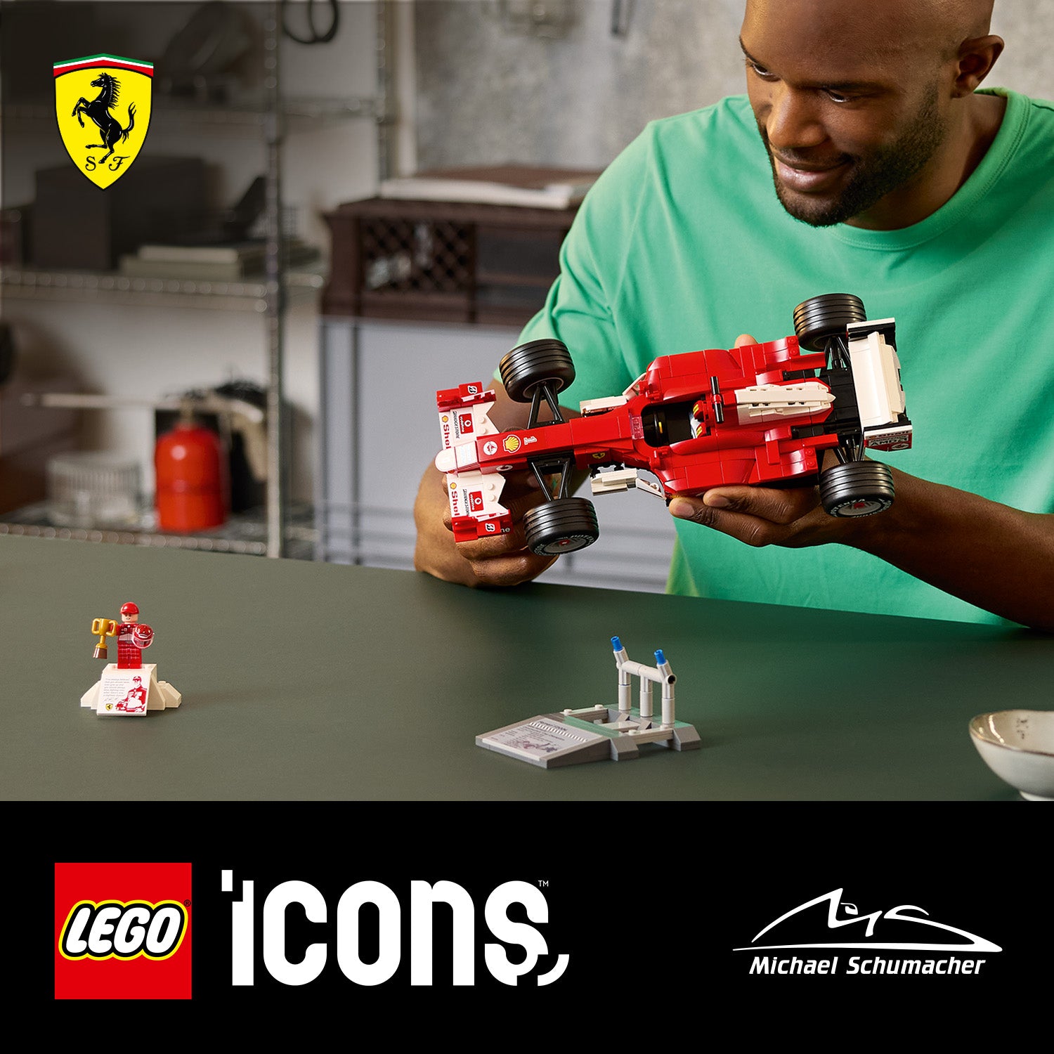 Person hält ein rotes LEGO Modell des Ferrari F2004 Formel-1-Rennwagens auf einem Tisch mit zwei kleinen LEGO Figuren und -Objekten, im Hintergrund das Ferrari-Logo und Logos von LEGO Icons und Michael Schumacher.