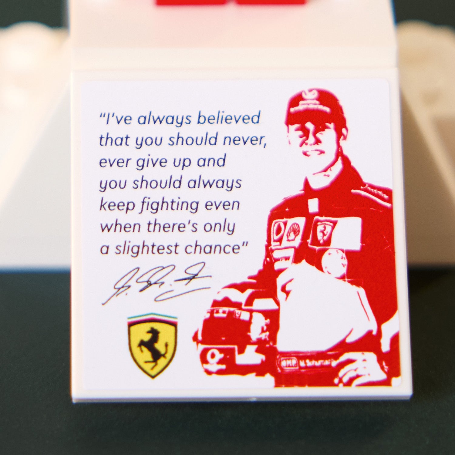 Rote LEGO Figur in Rennfahreranzug mit Ferrari-Logo neben Zitat und Ferrari-Wappen auf weißem Hintergrund