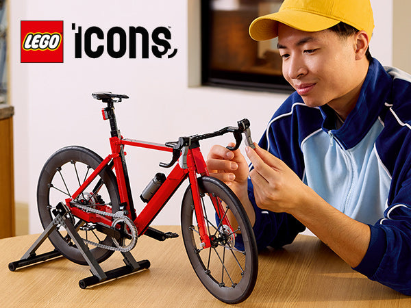 Rotes LEGO Rennrad-Modell auf einem schwarzen Ständer auf einem Tisch, daneben eine Person mit gelber Mütze und blau-weißem Pullover, die das Lenkrad hält, im Hintergrund das LEGO Icons Logo an der Wand.