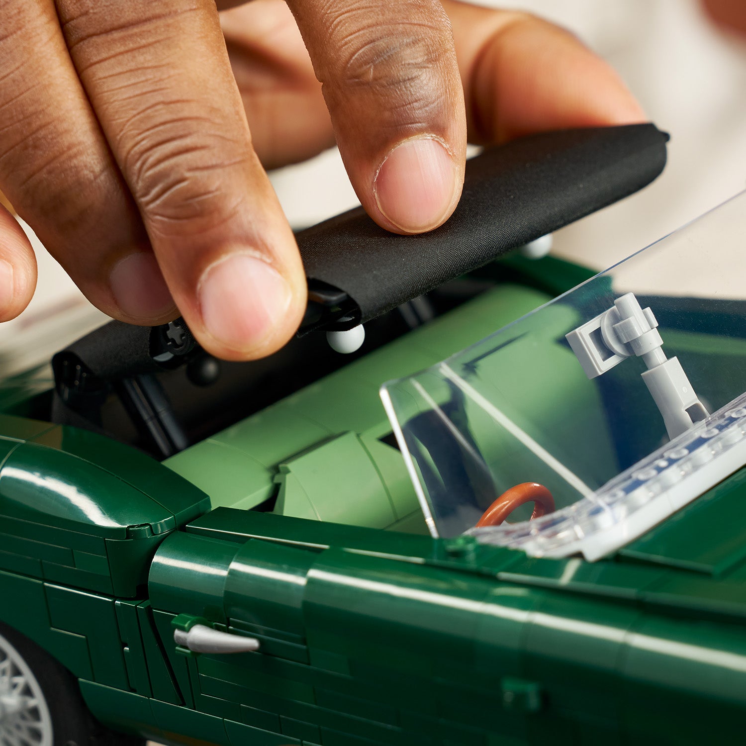 Hand hält schwarzen Spoiler über grünes LEGO Modell eines Jaguar E-Type mit sichtbarem Cockpit und Lenkrad