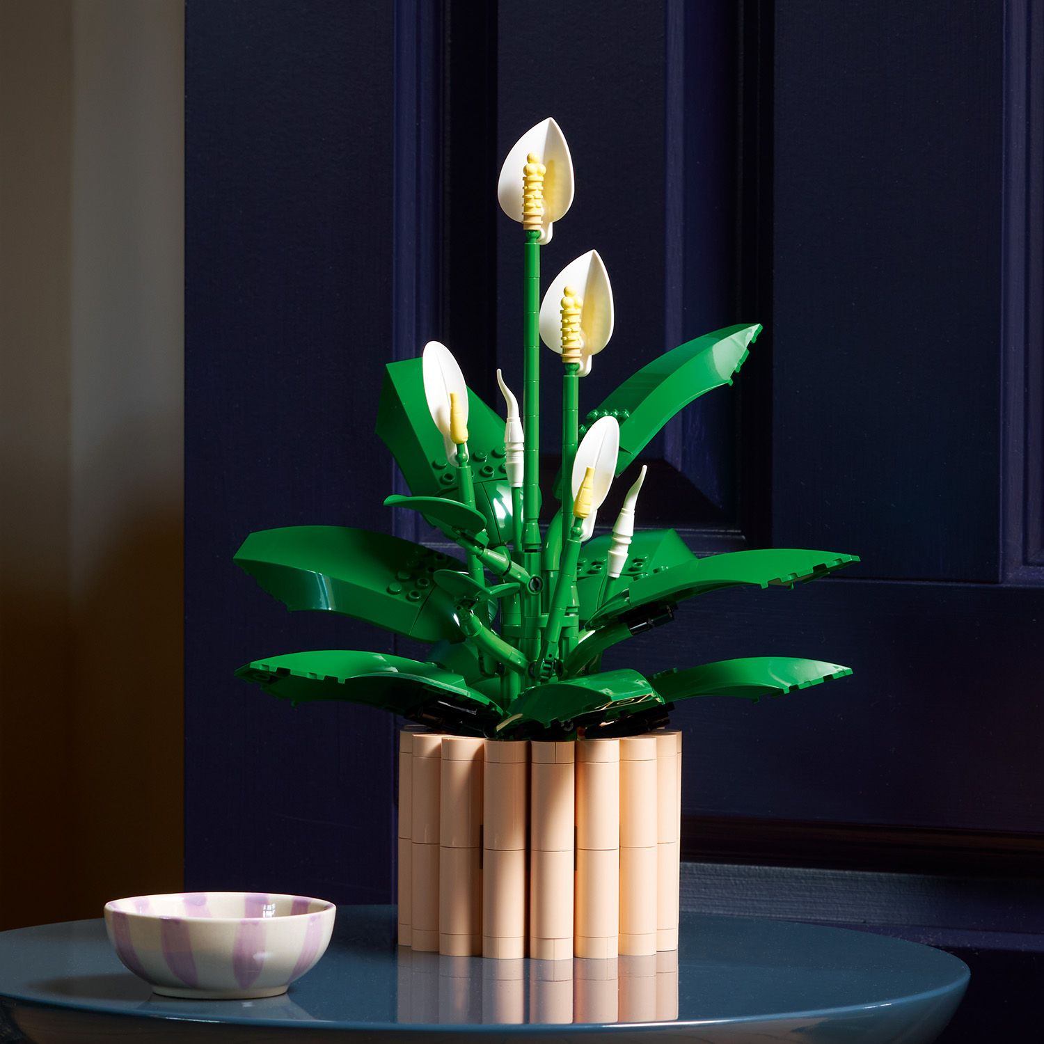 LEGO Modell einer Friedenslilie mit grünen Blättern und weißen Blüten in einem beigen Blumentopf aus LEGO Steinen auf einem runden blauen Tisch, daneben eine kleine Schale.