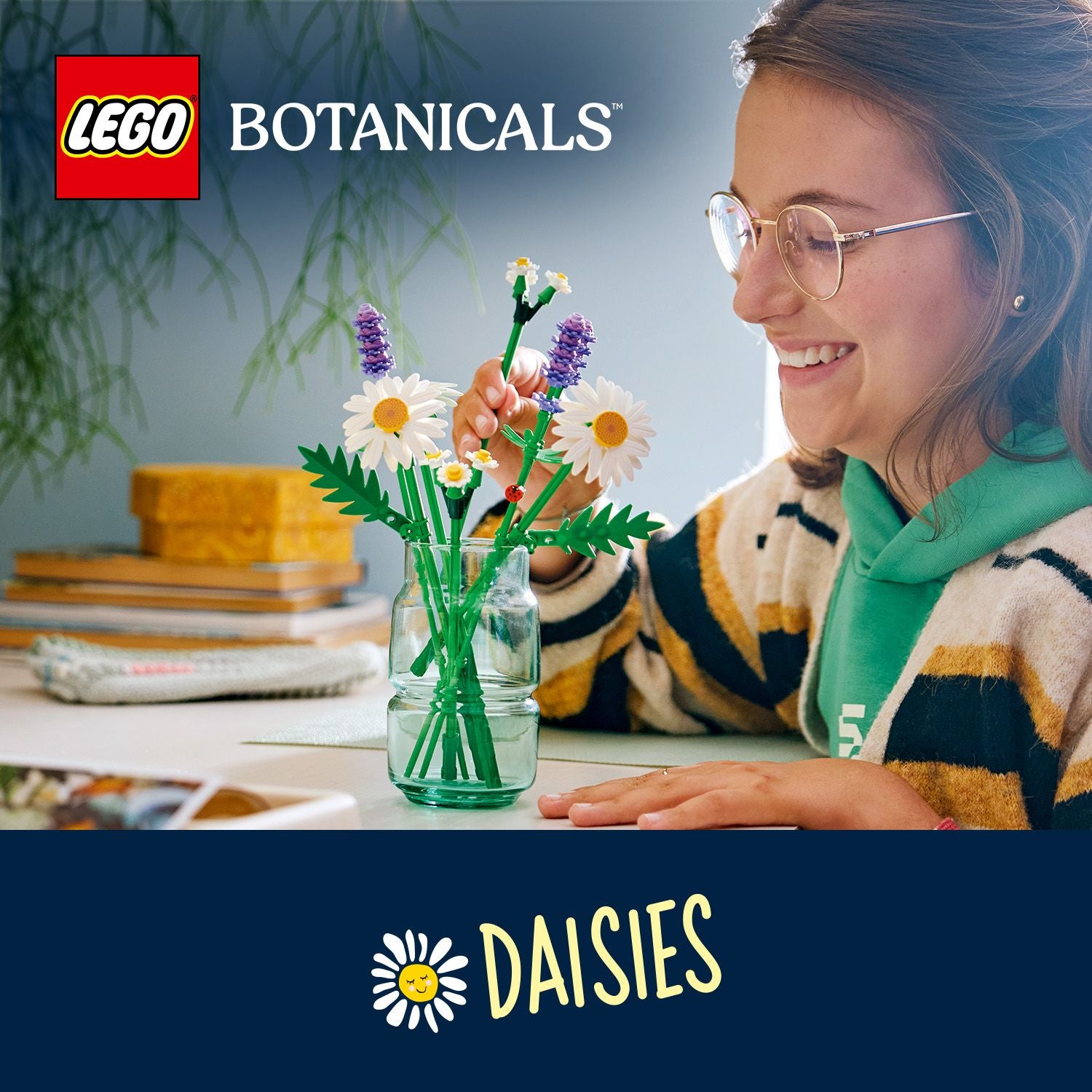 Frau mit grünem Hoodie und gestreiftem Pullover arrangiert LEGO Botanicals Gänseblümchen in einem Glas auf einem Tisch mit Büchern im Hintergrund.