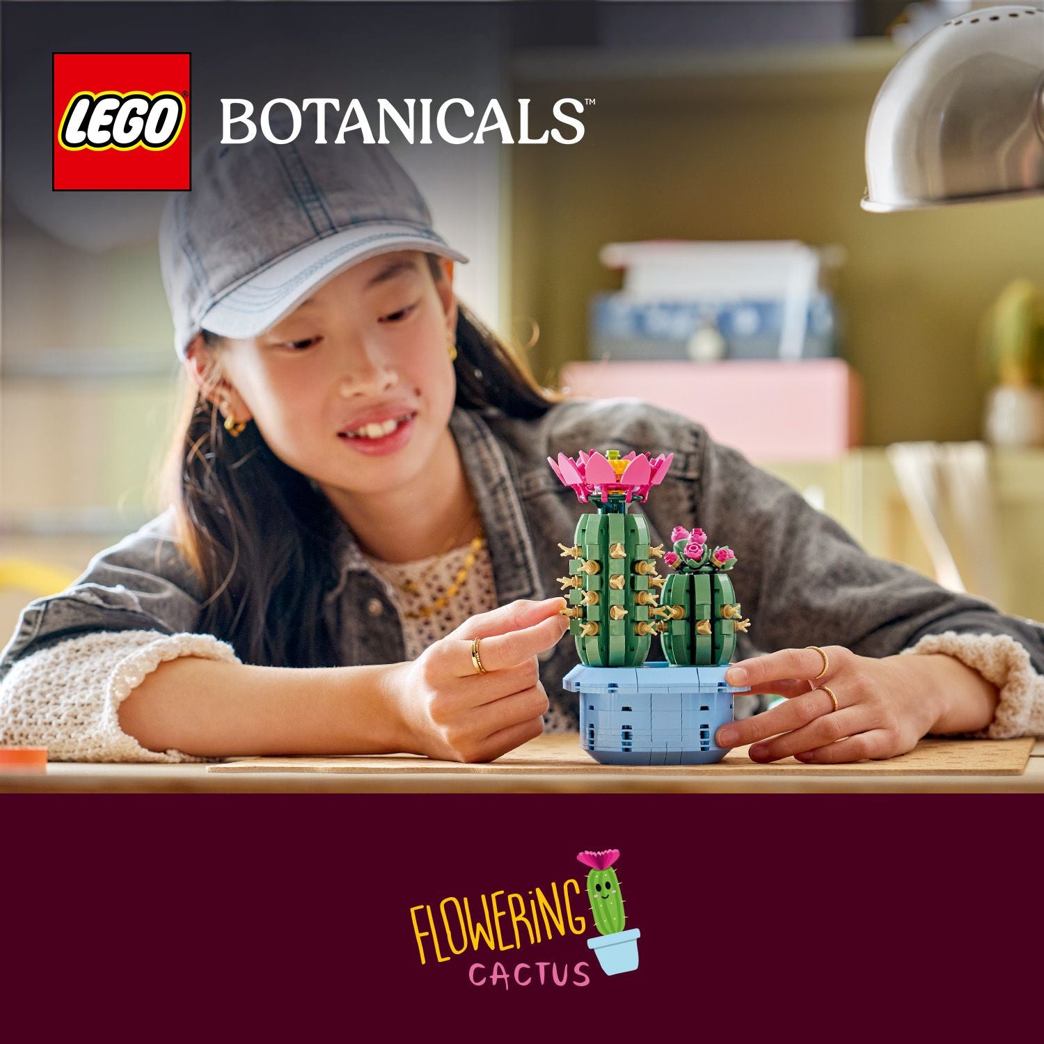 Eine Person hält auf einem Tisch zwei LEGO Botanicals-Kakteen in einem blauen Topf, einer mit pinken Blüten, vor einem unscharfen Hintergrund mit einem Lampenschirm.