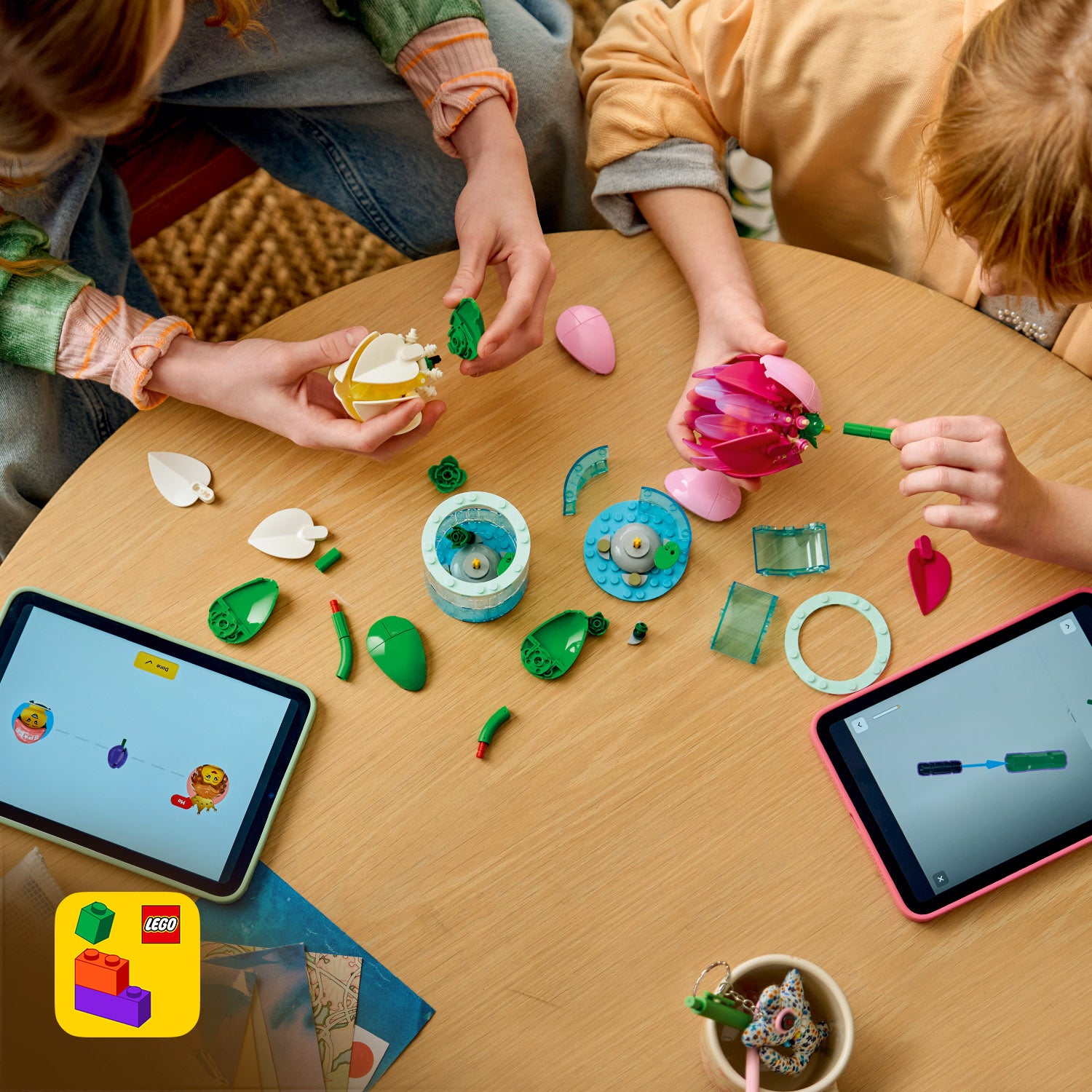 Zwei Personen bauen auf einem runden Holztisch mit LEGO Teilen eine Seerose, daneben liegen zwei Tablets mit LEGO Bauanleitungen.