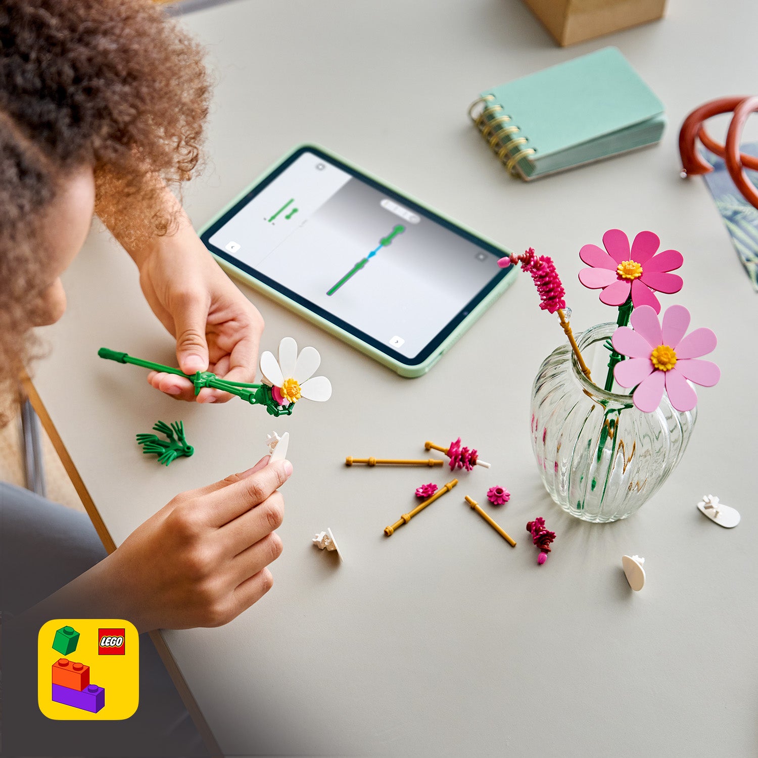 Kind baut mit LEGO Steinen eine Blume neben einem Glas mit LEGO Blumen auf einem Tisch mit Tablet und Notizbuch