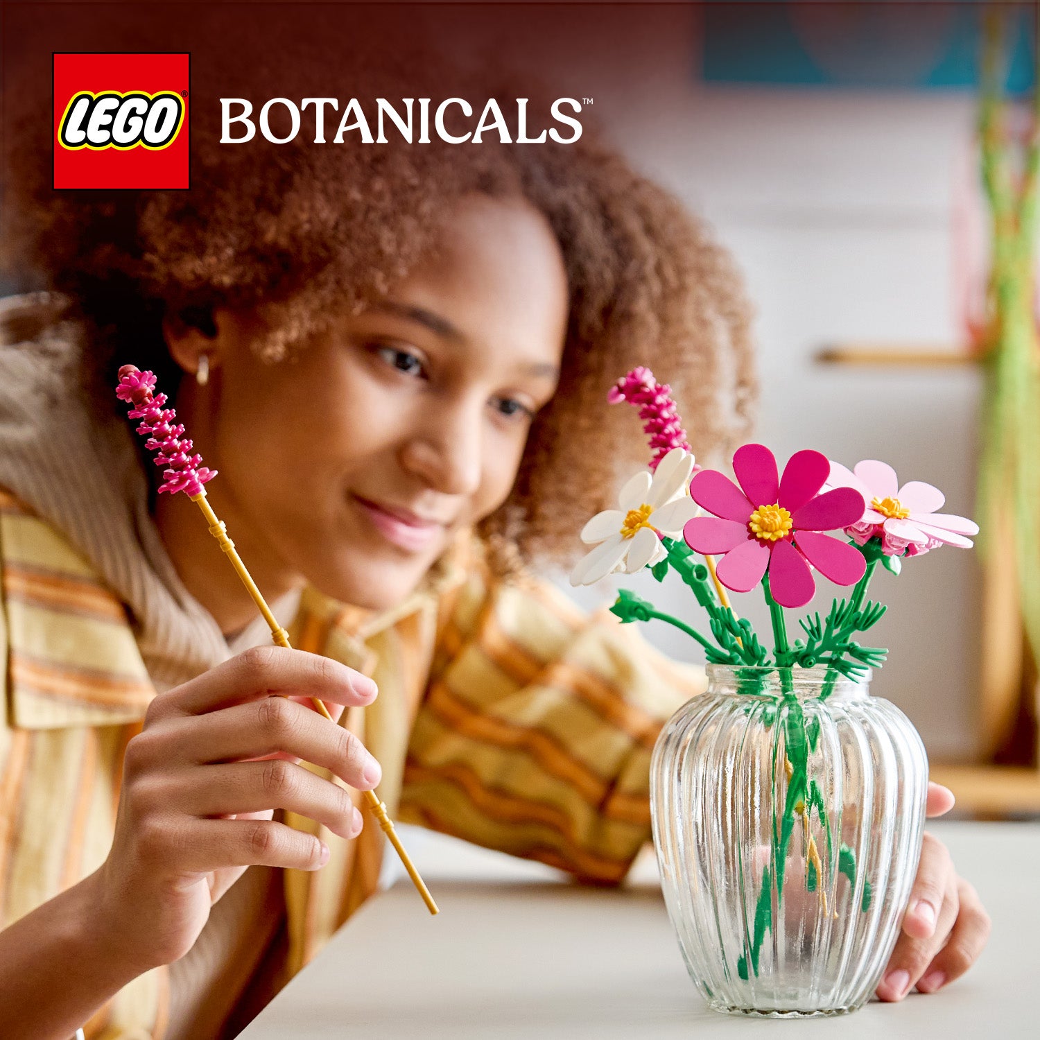 Junge Frau mit lockigem Haar hält zwei pinke LEGO-Blumenstiele über einer Glasvase mit LEGO Blumen in verschiedenen Farben