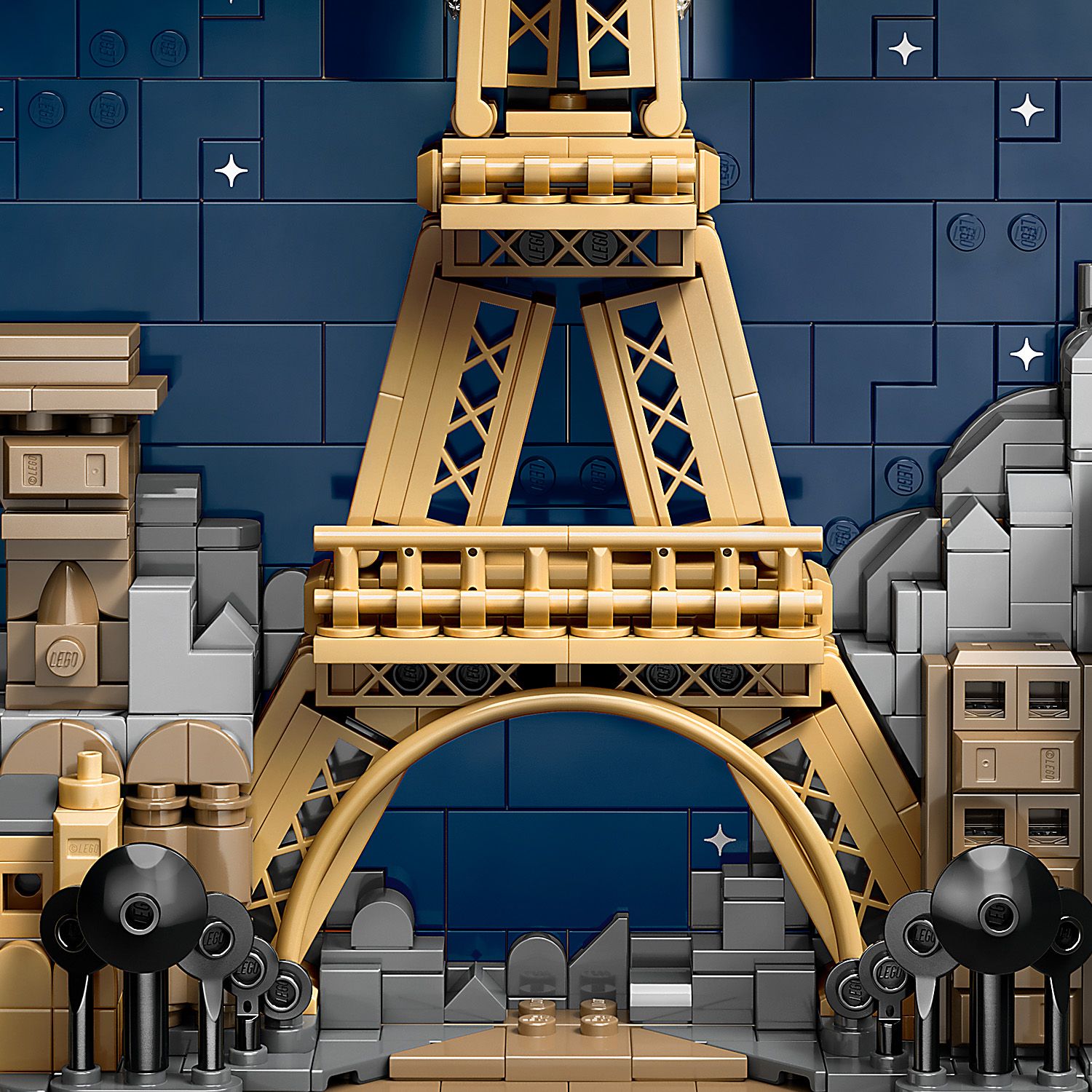 Detailaufnahme des goldfarbenen LEGO Modells des Eiffelturms vor einem dunkelblauen Hintergrund mit stilisierten Gebäuden und schwarzen Straßenlaternen im Vordergrund.