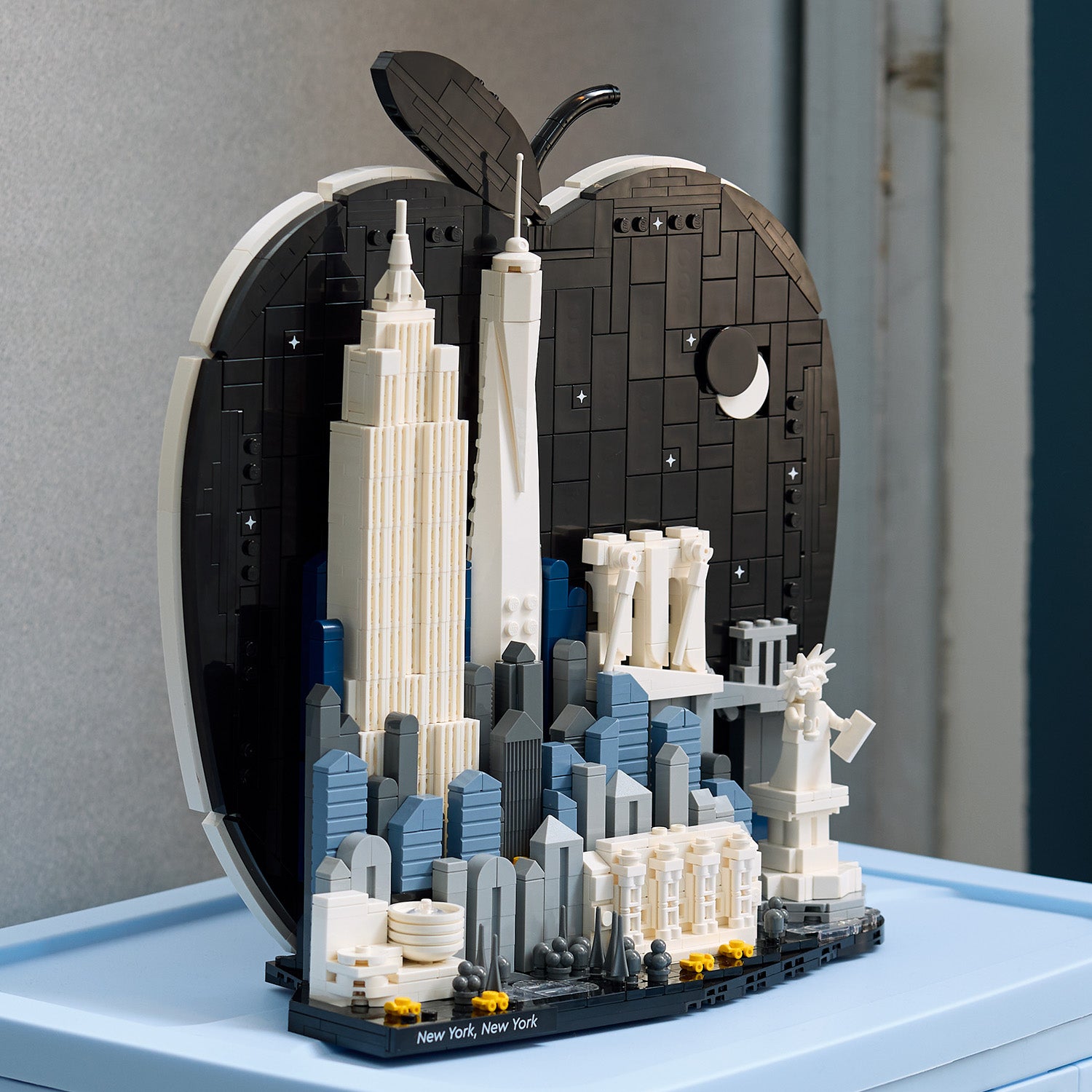 LEGO Modell der New Yorker Skyline mit Empire State Building, One World Trade Center, Freiheitsstatue und Brooklyn Bridge vor einer schwarzen Apfelform mit Mond und Sternen