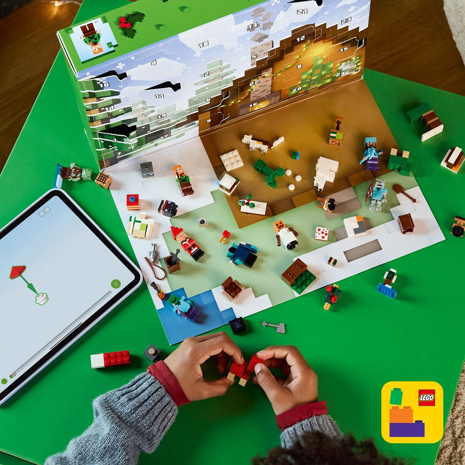 Aufgeklappter LEGO Minecraft Adventskalender 2025 mit kleinen LEGO-Figuren und Bauteilen auf grünem Tisch, Hände bauen ein rotes LEGO Teil, Tablet mit LEGO Anleitung, LEGO Logo unten rechts.