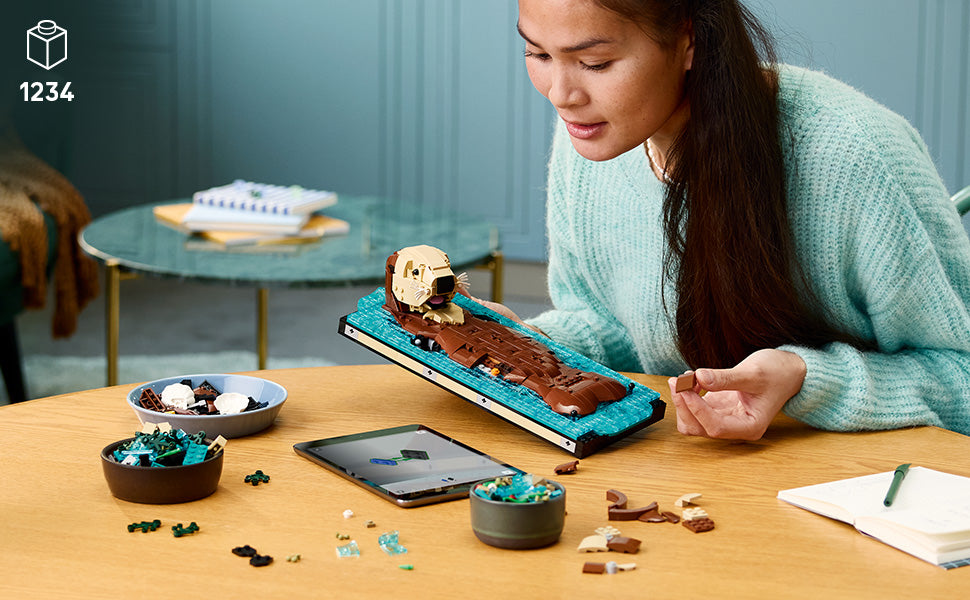 Eine Person mit langem Haar hält ein LEGO Modell eines schwimmenden Otters auf einer blauen Grundplatte auf einem Holztisch mit verstreuten LEGO Teilen und einem Tablet im Hintergrund