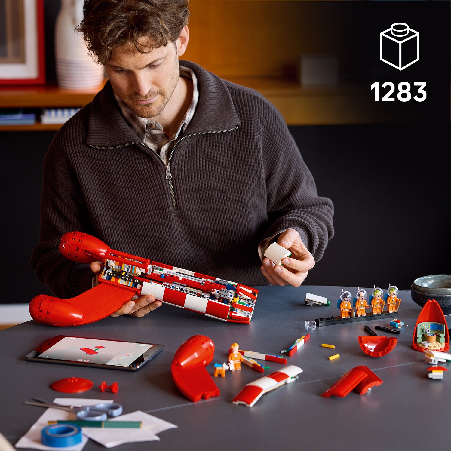 Mann baut eine rote LEGO Rakete mit mehreren orangefarbenen Astronautenfiguren auf einem Tisch mit Tablet und Bauanleitung