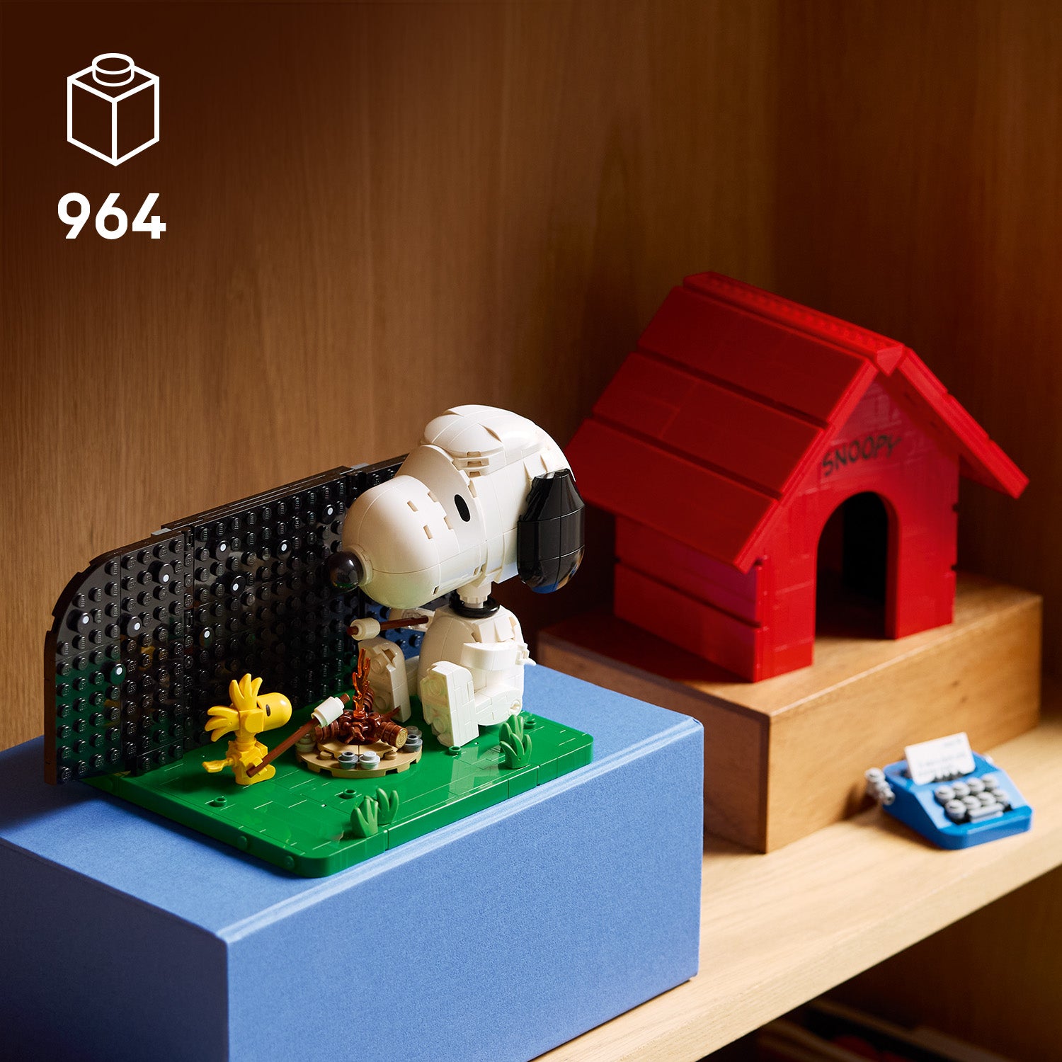 LEGO Modell von Snoopy mit Woodstock vor einem Lagerfeuer auf grünem Untergrund neben einer roten Hundehütte mit der Aufschrift 'SNOOPY' und einer kleinen blauen Schreibmaschine auf einem Holzregal