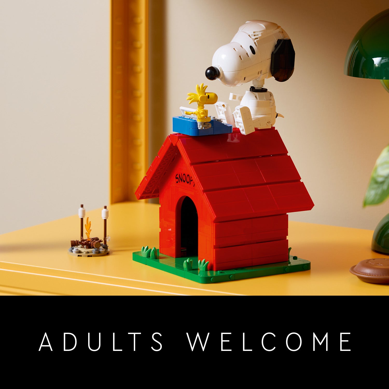 LEGO Modell der roten Hundehütte mit der Aufschrift 'SNOOPY', darauf sitzend die Figuren Snoopy und Woodstock aus den Peanuts, daneben ein kleines Lagerfeuer aus LEGO Steinen auf einem gelben Tisch.