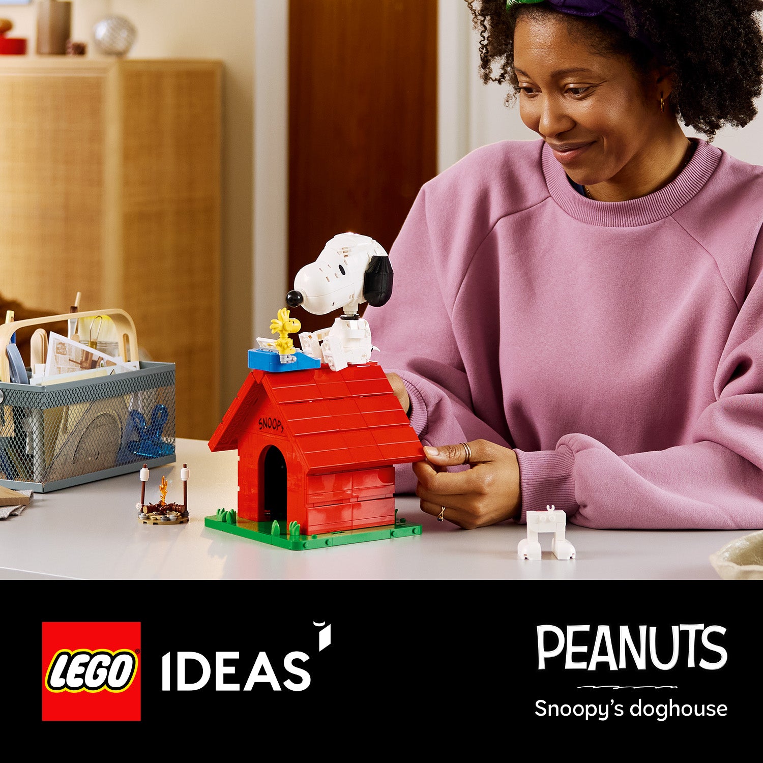 Eine Person in rosa Pullover sitzt an einem Tisch und hält ein rotes LEGO Modell von Snoopys Hundehütte mit der Figur Snoopy und dem Vogel Woodstock darauf.