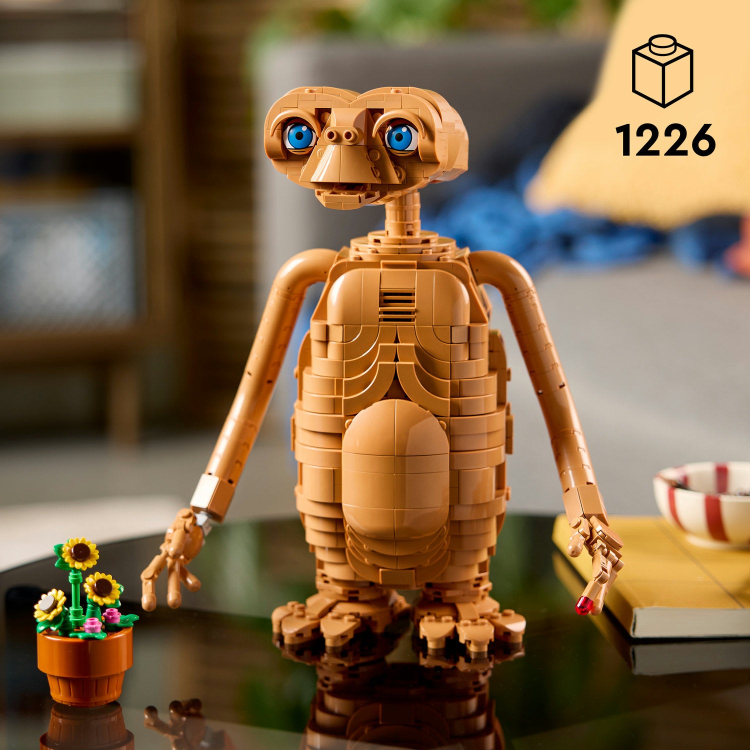 Braune LEGO Figur von E.T. mit großen blauen Augen, ausgestreckten Armen und einem leuchtenden Finger, stehend auf einem Tisch neben einem Blumentopf mit Sonnenblumen und einem Buch.