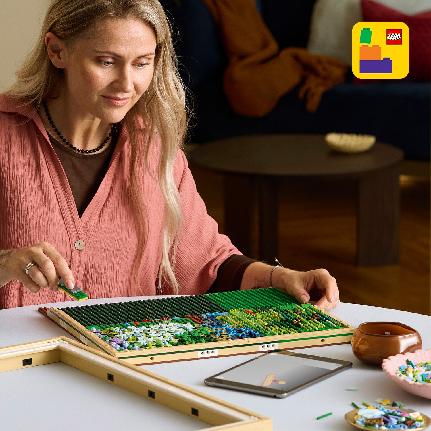 Frau mit langem blonden Haar baut an einem LEGO Art-Mosaik mit grünen und blauen Steinen auf einem weißen Tisch