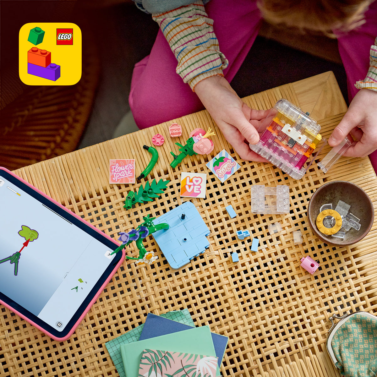 Kind baut mit LEGO Steinen eine Parfümflasche mit Blumendekoration auf einem geflochtenen Holztisch neben einem Tablet mit LEGO Bauanleitung