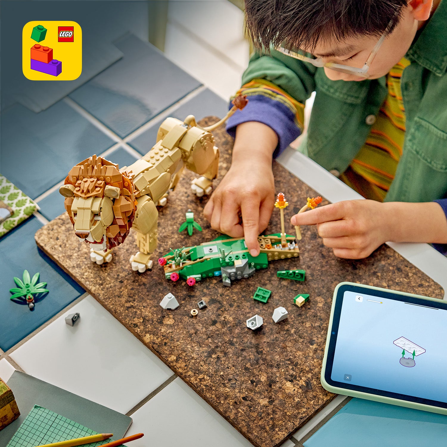 Kind baut mit LEGO Steinen einen Löwen und ein Krokodil auf einer Korkplatte, daneben ein Tablet mit Bauanleitung und verstreute LEGO Teile