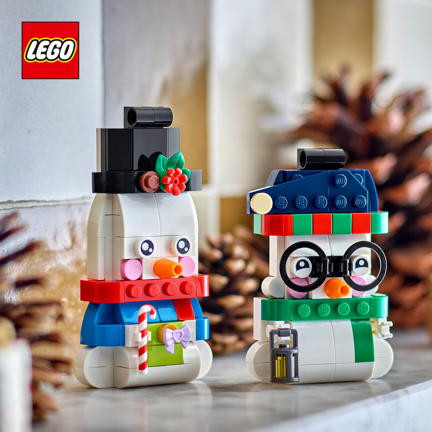 Zwei LEGO Schneemänner mit bunten Mützen, einer mit Zuckerstange und der andere mit Laterne, vor unscharfem Hintergrund mit Tannenzapfen
