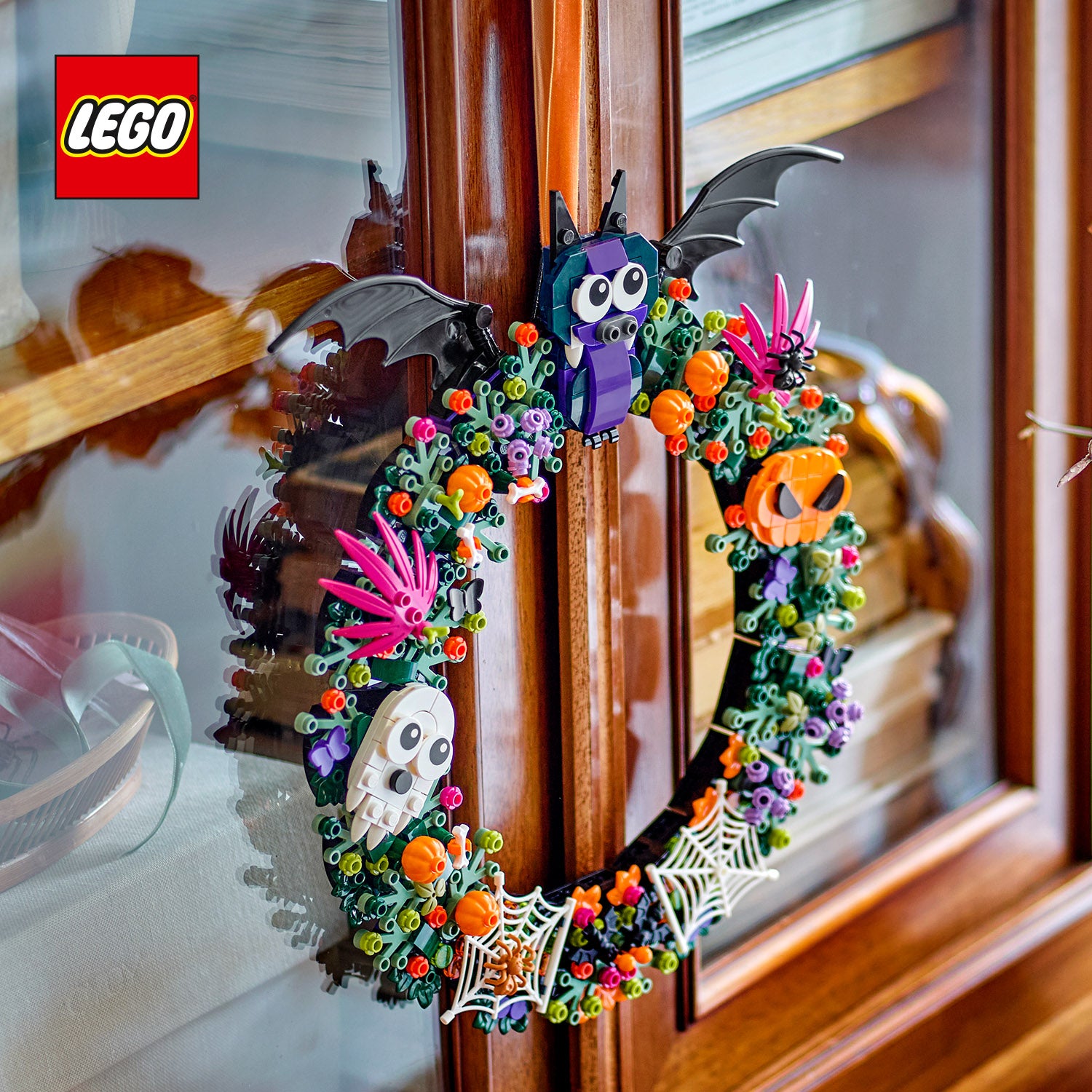Halloweenkranz aus LEGO Steinen mit Fledermaus, Totenkopf, Kürbis, Spinnennetzen und bunten Blüten an einer Holztür hängend