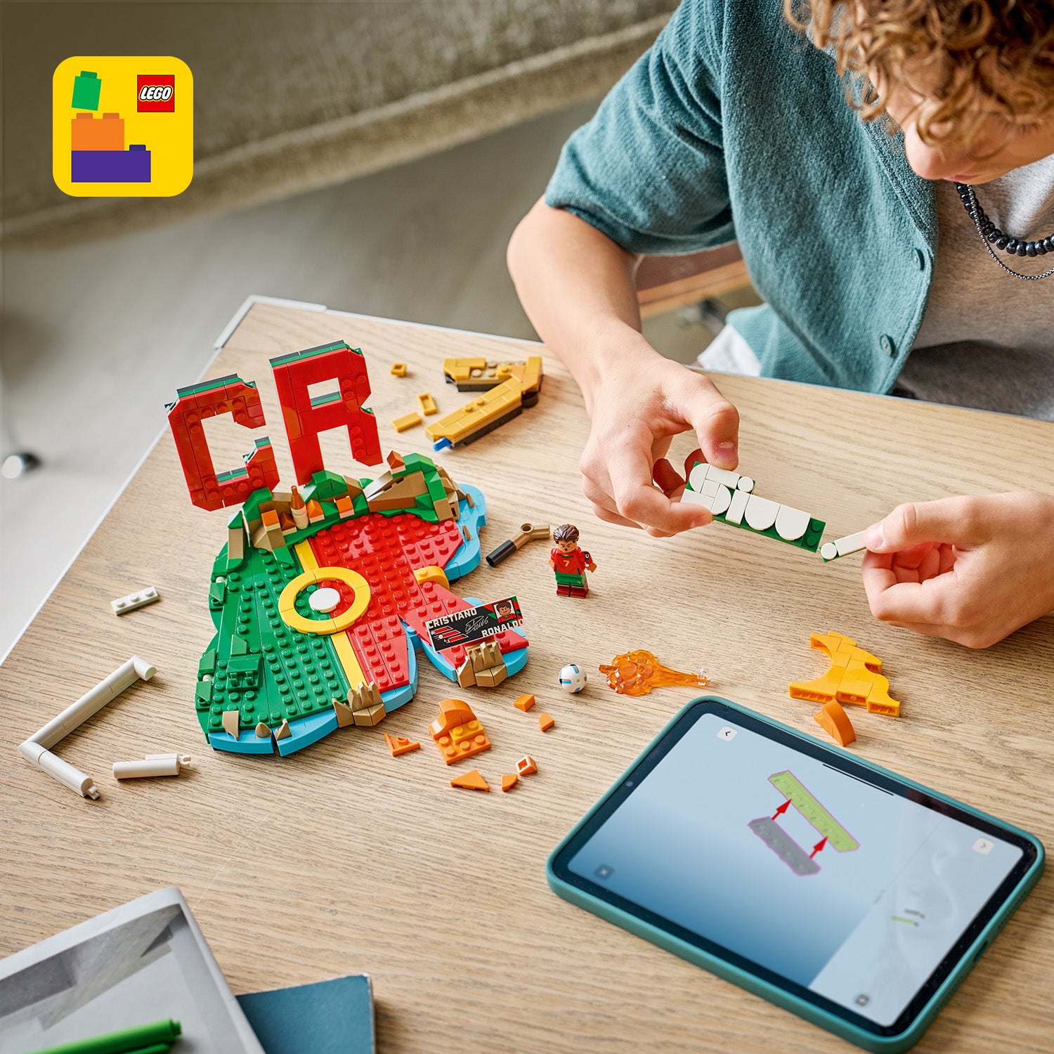 Person baut LEGO Set mit Fußballfeld, roten Buchstaben "CR" und Minifigur, daneben Tablet mit Bauanleitung