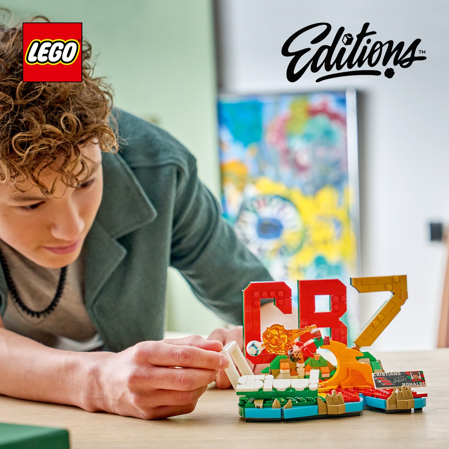 LEGO Edition mit dem Schriftzug 'CR7' aus LEGO Steinen und einer kleinen LEGO Figur auf einem Tisch, im Hintergrund unscharfe bunte Wanddekoration, eine Person mit lockigem Haar und grünem Oberteil beugt sich über den Tisch