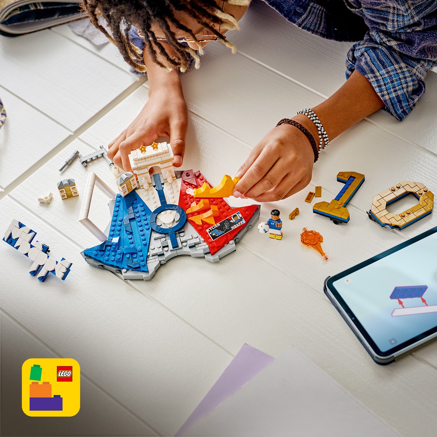 Hände bauen mit LEGO Steinen ein rundes Modell mit blauen, roten und grauen Segmenten und einer kleinen LEGO Figur auf einem weißen Tisch neben einem Tablet mit LEGO Anleitung.