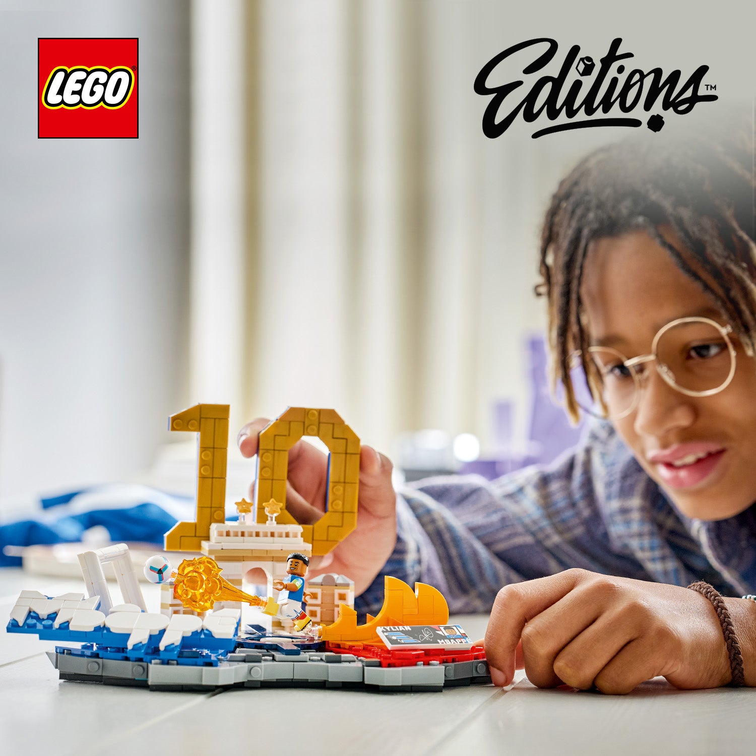 Kind spielt mit LEGO Set, das eine kleine Bühne mit der Zahl 10 und eine Minifigur zeigt, im Hintergrund unscharf ein Raum mit Vorhängen