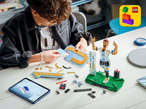 Junge baut am Tisch eine LEGO Figur von Lionel Messi mit Fußballtrikot und hebt eine LEGO Platte über einem Tablet mit Bauanleitung.