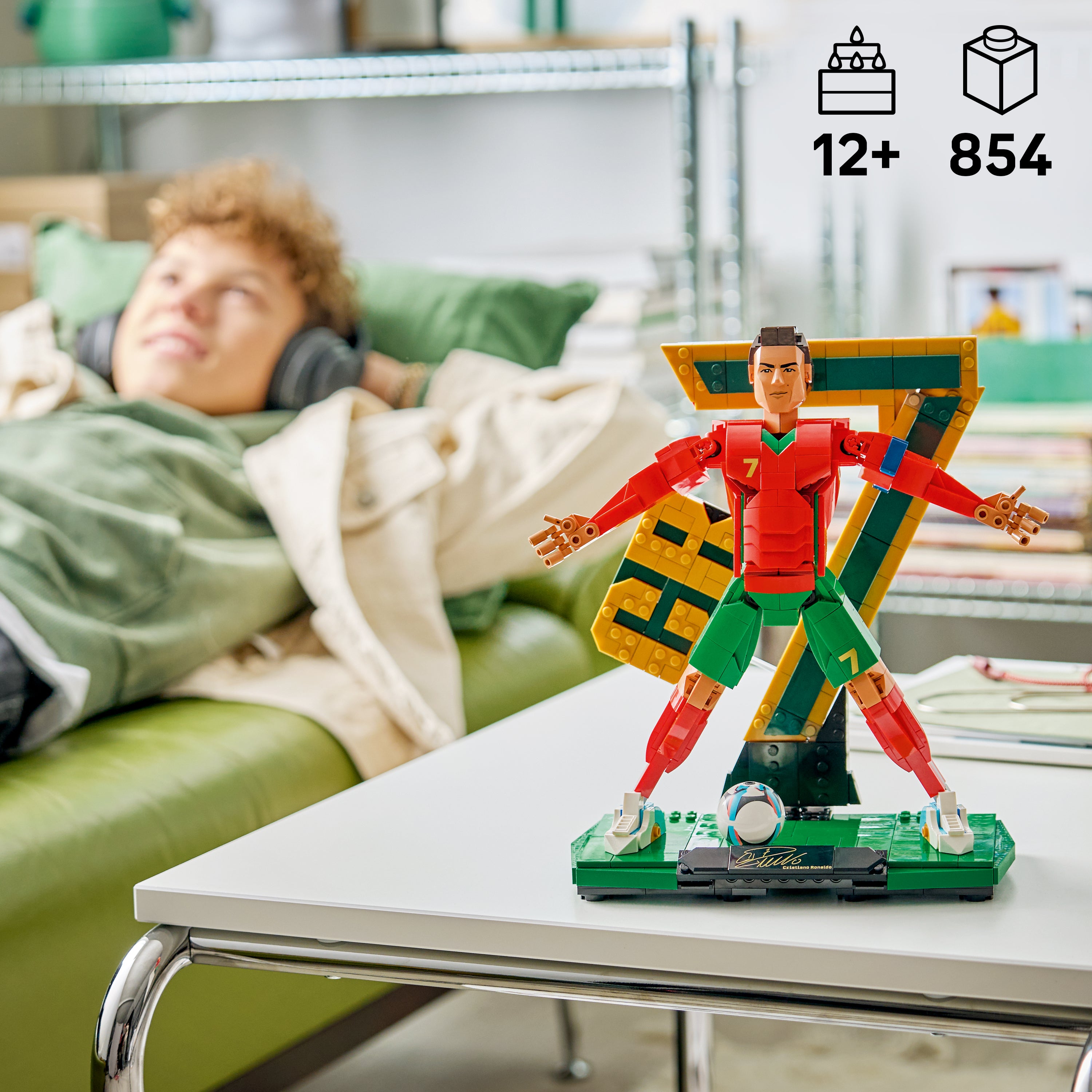 LEGO Figur von Cristiano Ronaldo in rotem Trikot und grüner Hose mit ausgestreckten Armen auf einem weißen Tisch vor einem Sofa mit einer Person im Hintergrund