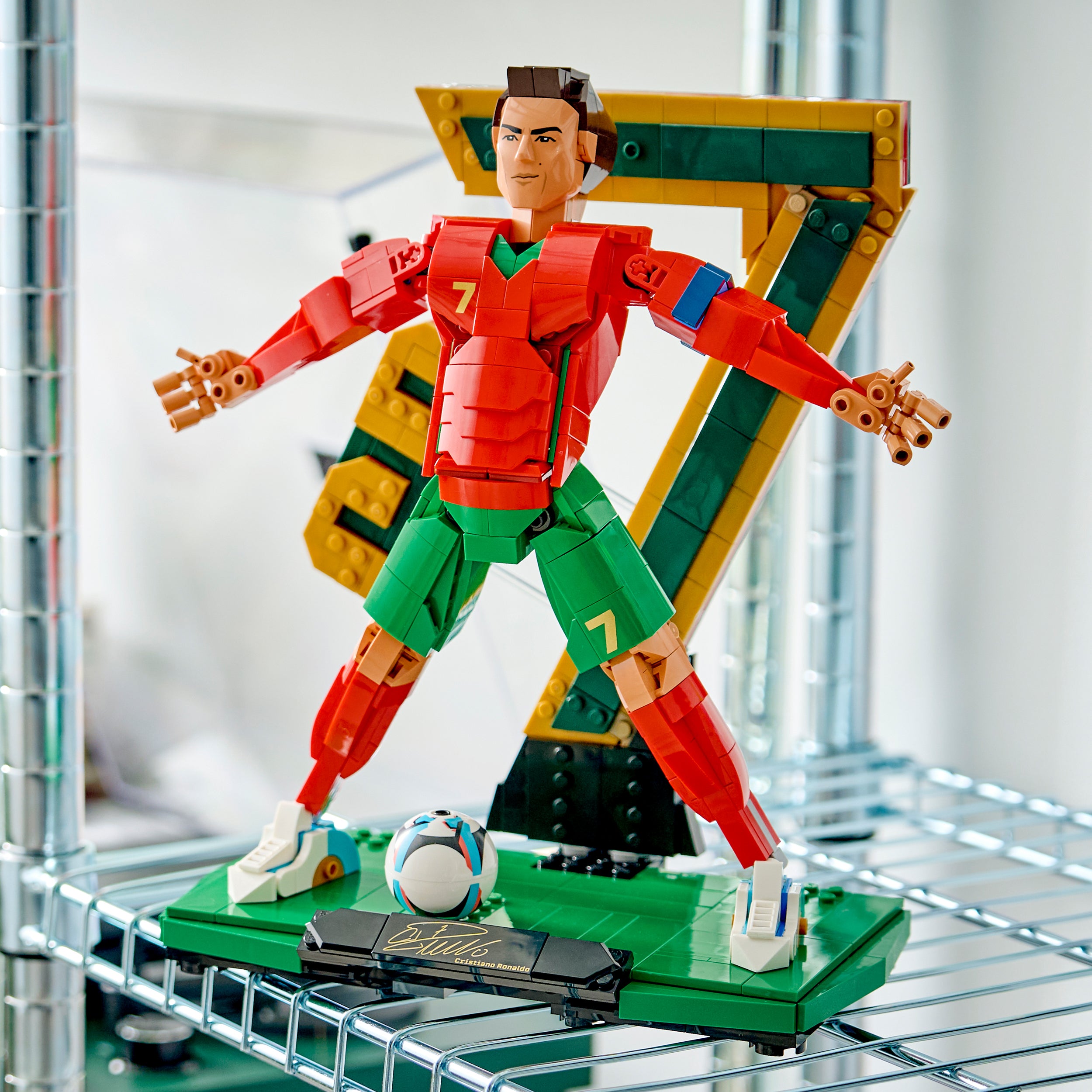 LEGO Figur eines Fußballspielers in rotem Trikot mit der Nummer 7 und grüner Hose steht mit gespreizten Armen auf grünem Sockel vor einer großen goldenen Zahl 7 aus LEGO Steinen