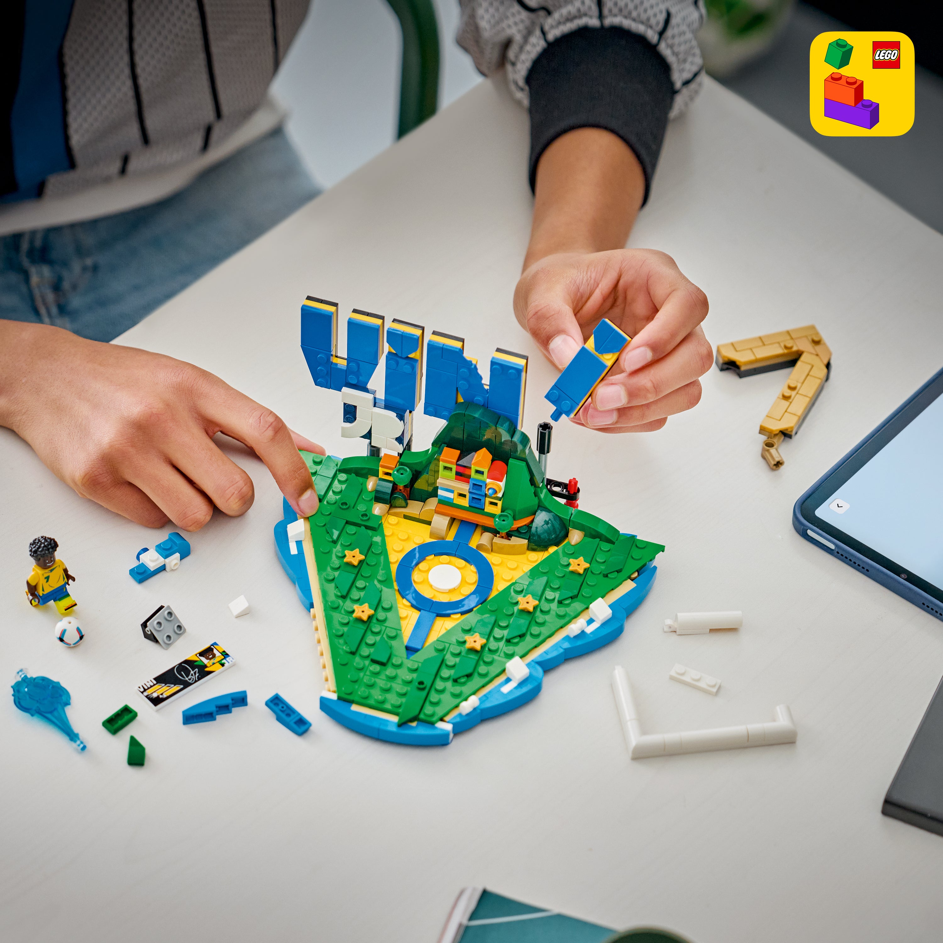 Kinderhände bauen mit LEGO-Steinen ein grünes, dreieckiges Spielfeld mit dem Schriftzug 'VINI' im Hintergrund