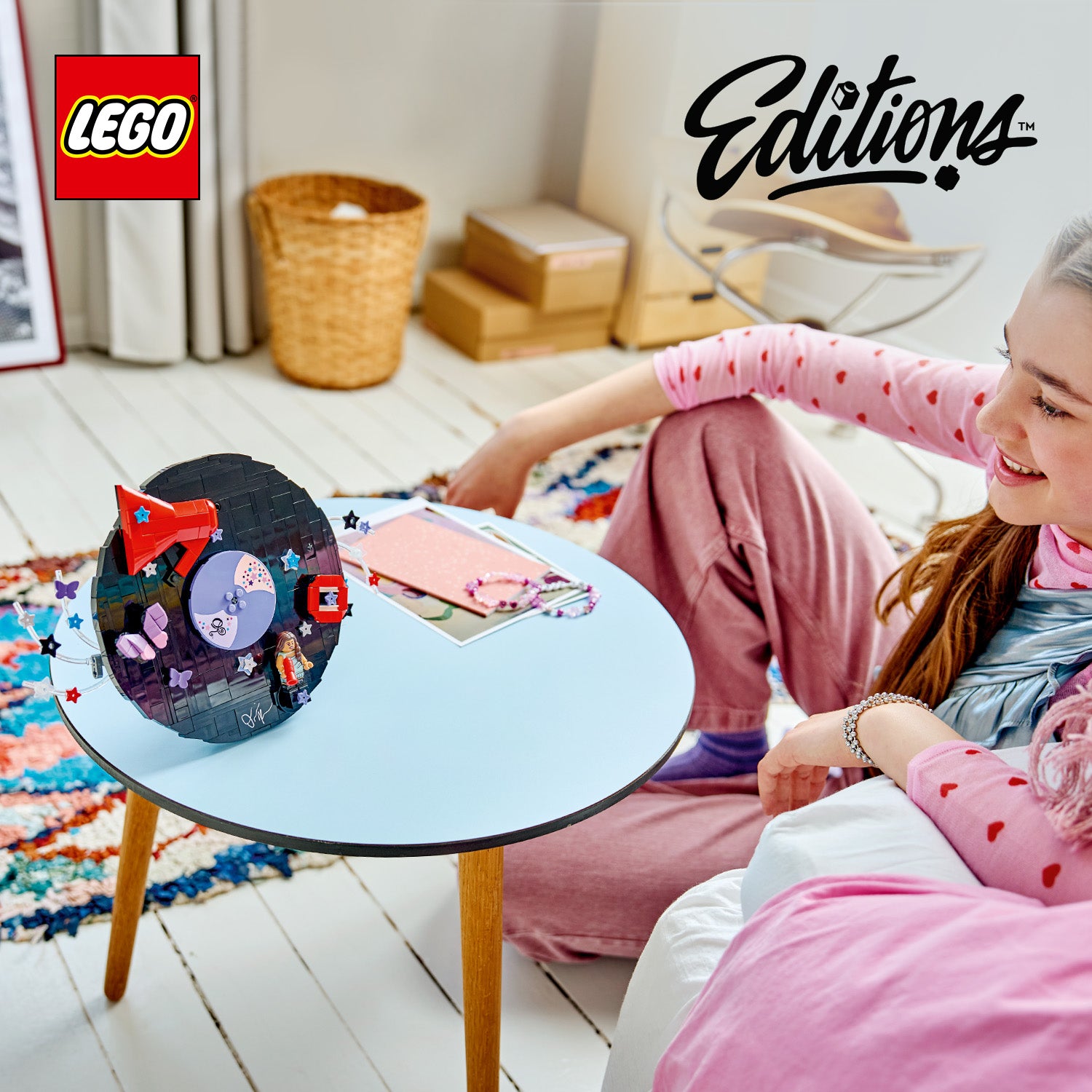 Ein runder, schwarzer LEGO Schallplatten-Bausatz mit roten und lilafarbenen Details steht auf einem runden weißen Tisch vor einer jungen Frau mit rosa Haaren, die auf einem weißen Sofa sitzt.