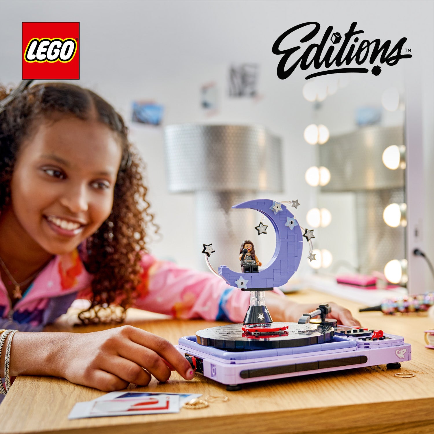 Ein Mädchen mit lockigem Haar spielt an einem Tisch mit einem lila LEGO Set, das eine Mondform mit einer Minifigur und Sternen zeigt, auf einem Plattenspieler-ähnlichen Sockel.