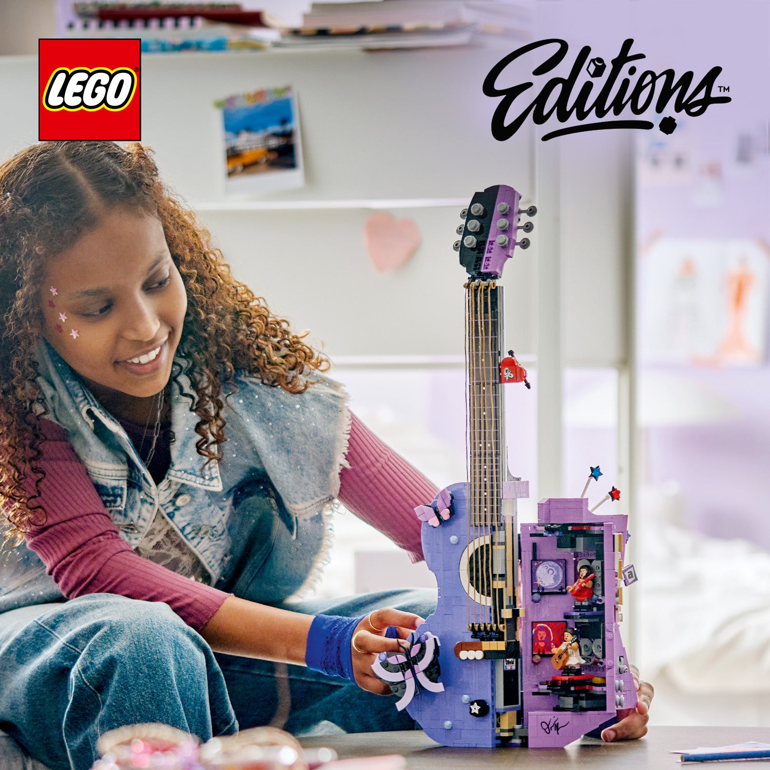 Ein Mädchen mit lockigem Haar sitzt auf dem Boden und hält eine lila LEGO Gitarre aus der LEGO Editions Serie, die detaillierte Bauelemente und kleine Figuren zeigt