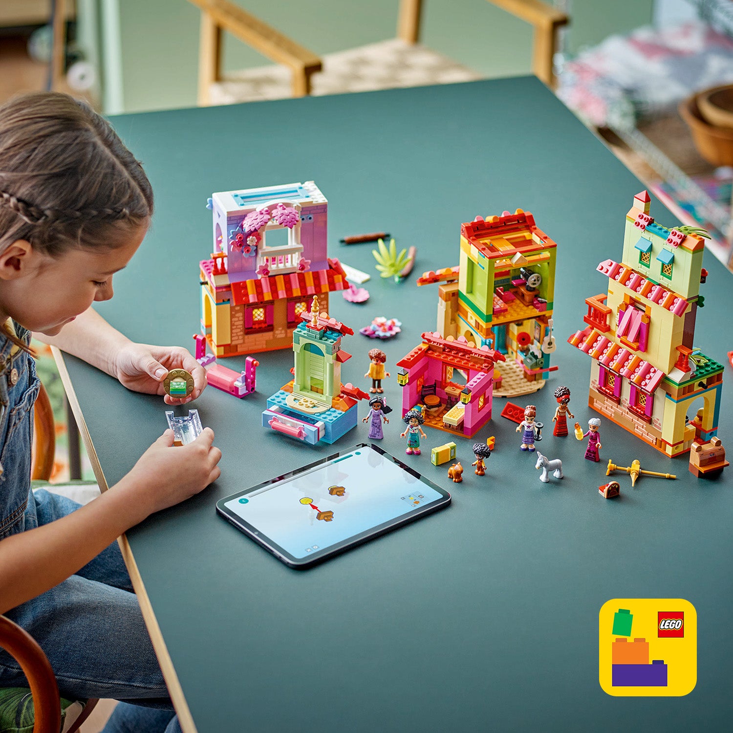 Kind spielt mit LEGO Set "Das magische Haus der Madrigals" mit mehreren bunten Gebäuden, Figuren und Tieren auf einem Tisch, daneben ein Tablet mit Bauanleitung.