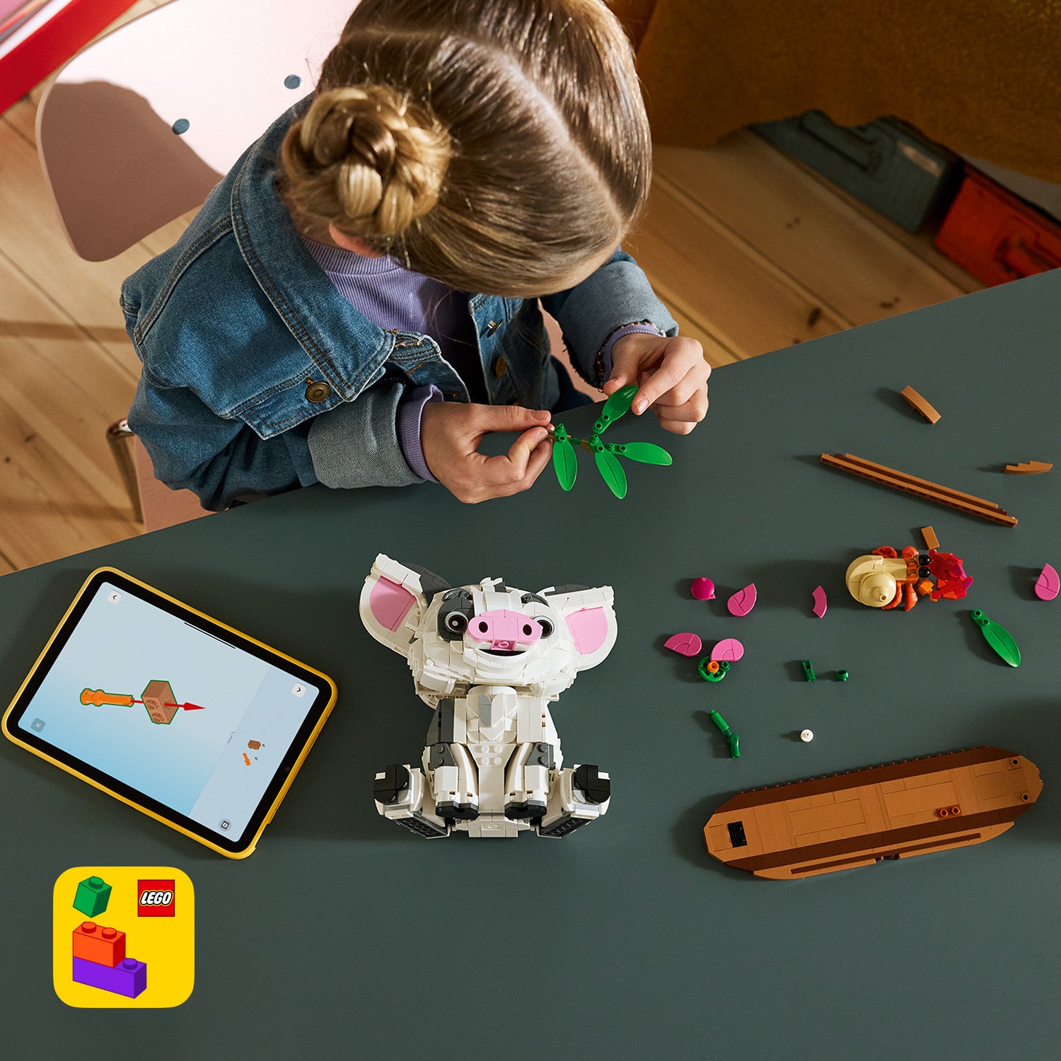 Kind mit zusammengebundenem Haar baut am Tisch eine grüne LEGO Pflanze neben einem LEGO Modell des Schweins Pua und einem Tablet mit LEGO App