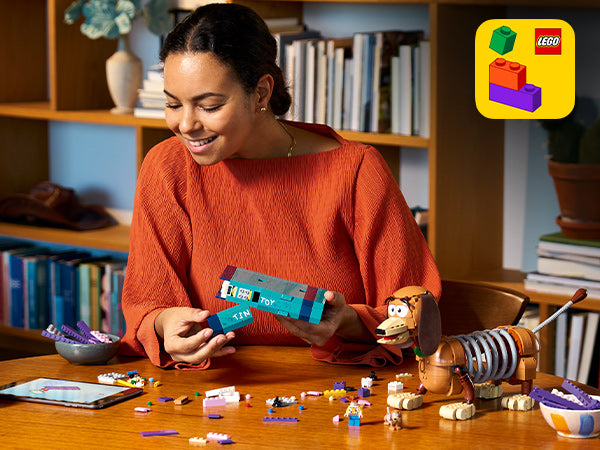 Frau in orangefarbenem Pullover sitzt an einem Holztisch und hält ein blaues LEGO Modell mit der Aufschrift 'TIN TOY', daneben steht ein LEGO Modell der Disney-Figur Slinky Dog mit ausziehbarem Metallkörper.