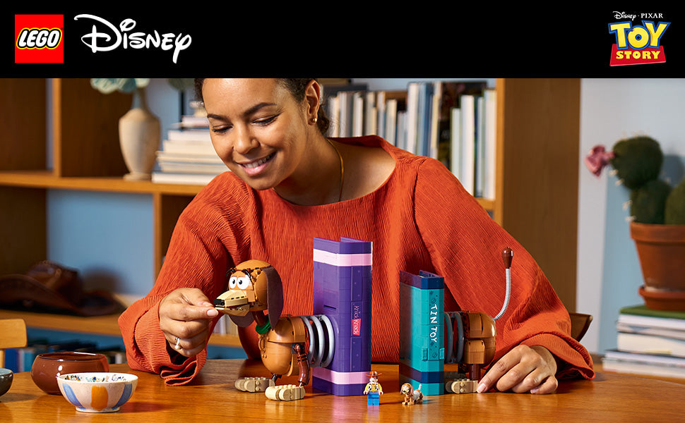 Person in orangerotem Pullover sitzt an Holztisch und hält LEGO Spielzeugfigur von Slinky Dog aus Toy Story neben zwei bunten LEGO Buchstützen