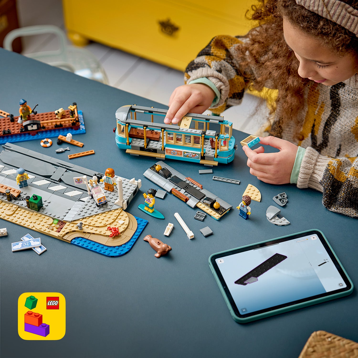 Kind baut mit LEGO Steinen eine klassische Strandbahn auf einem Tisch, umgeben von LEGO Figuren und Zubehör, daneben ein Tablet mit LEGO Bauanleitung