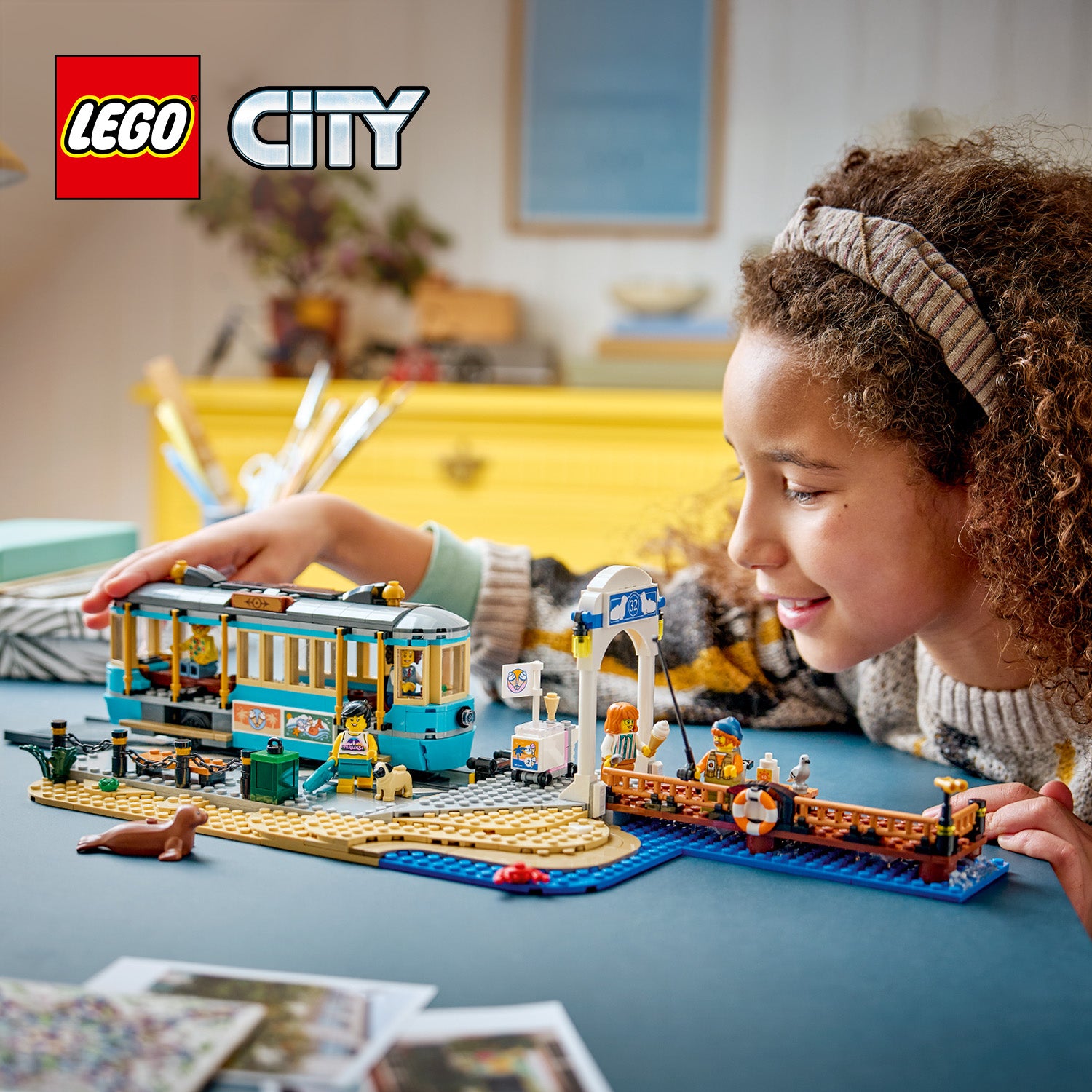Kind spielt mit LEGO City Set einer klassischen Strandbahn mit blauer Straßenbahn, Anlegestelle und Figuren auf einem Tisch