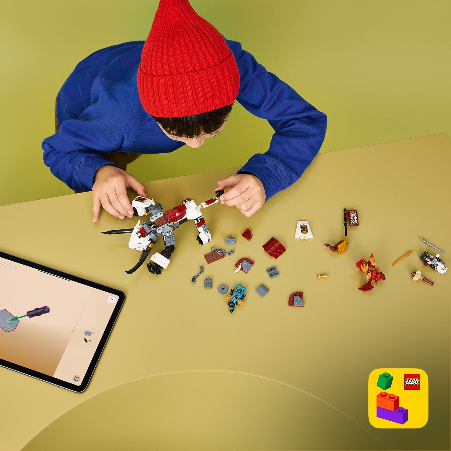 Kind mit roter Mütze und blauem Pullover baut LEGO Drachentier Grimtak mit verstreuten LEGO Teilen und Tablet mit Bauanleitung auf gelbem Untergrund