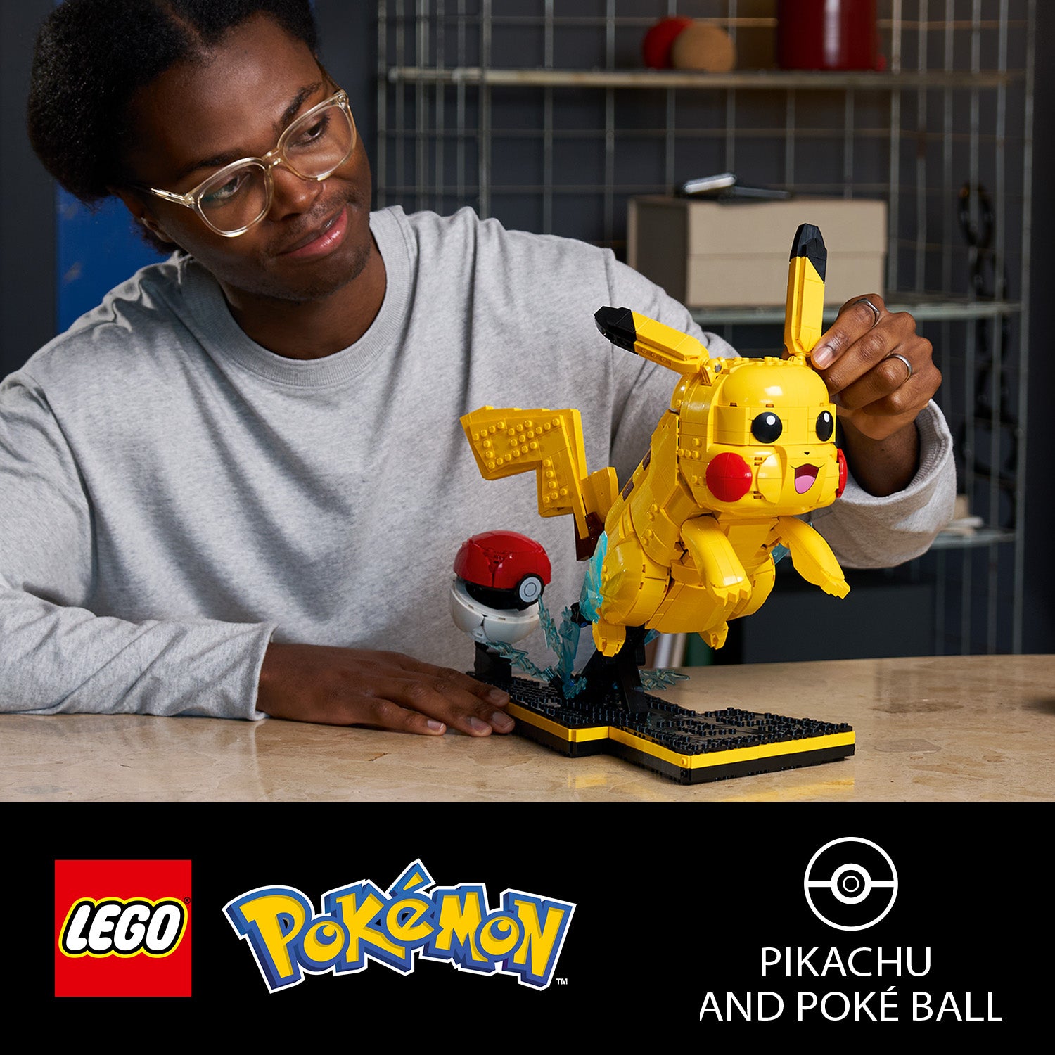 Eine LEGO Figur von Pikachu mit einem Pokéball auf einem schwarzen Sockel, gehalten von einer Person mit grauem Langarmshirt an einem Holztisch.