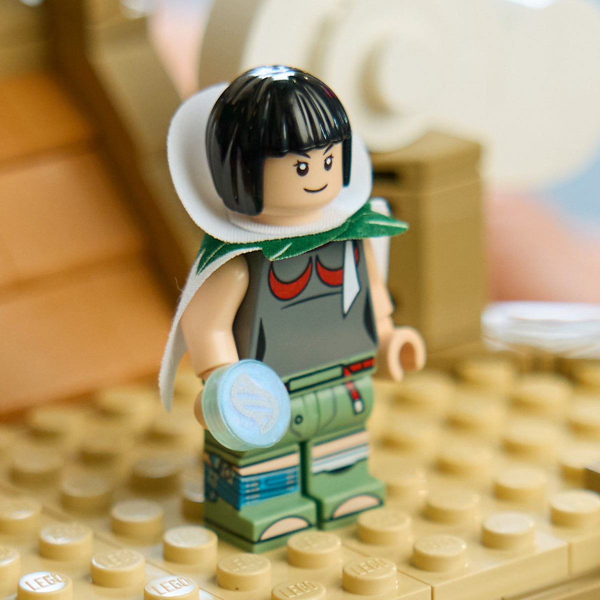LEGO Figur mit schwarzem Bob, grauem Oberteil, grüner Hose und weißem Umhang steht auf beige LEGO Platte